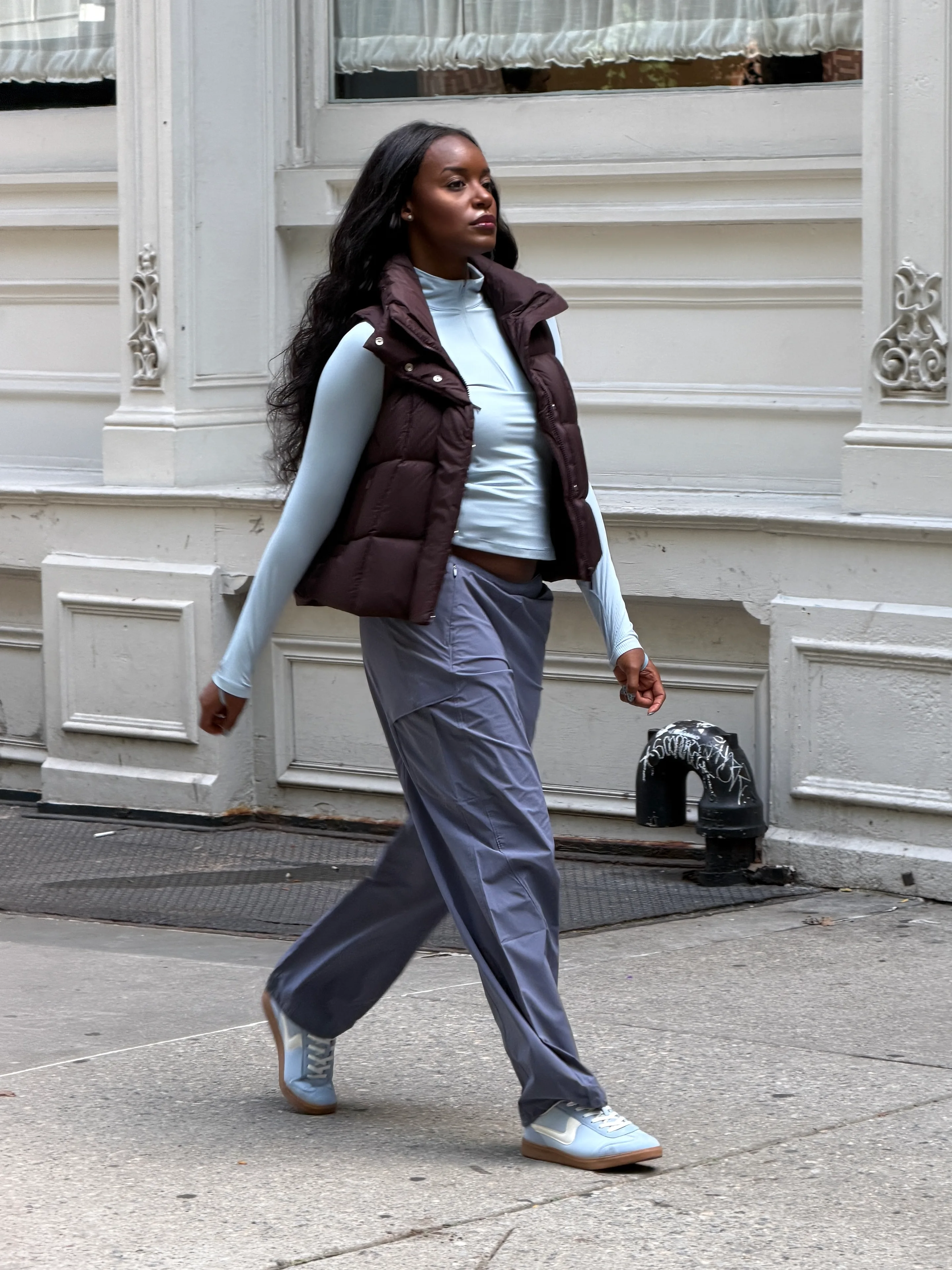 Brown puffer vest over a light blue turtleneck with blue wide-leg pants and sneakers.