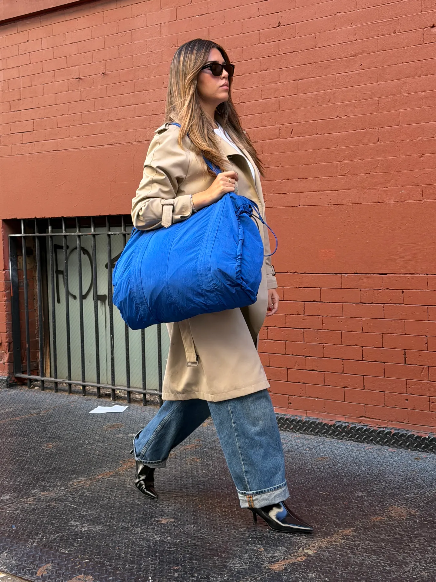 Beige trench coat with blue duffle bag, rolled-cuff jeans, black heels, and sunglasses.