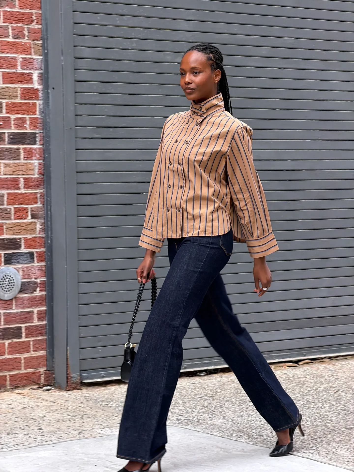 Tan striped button-up shirt, dark wash jeans, black heeled mules, and a black handbag.