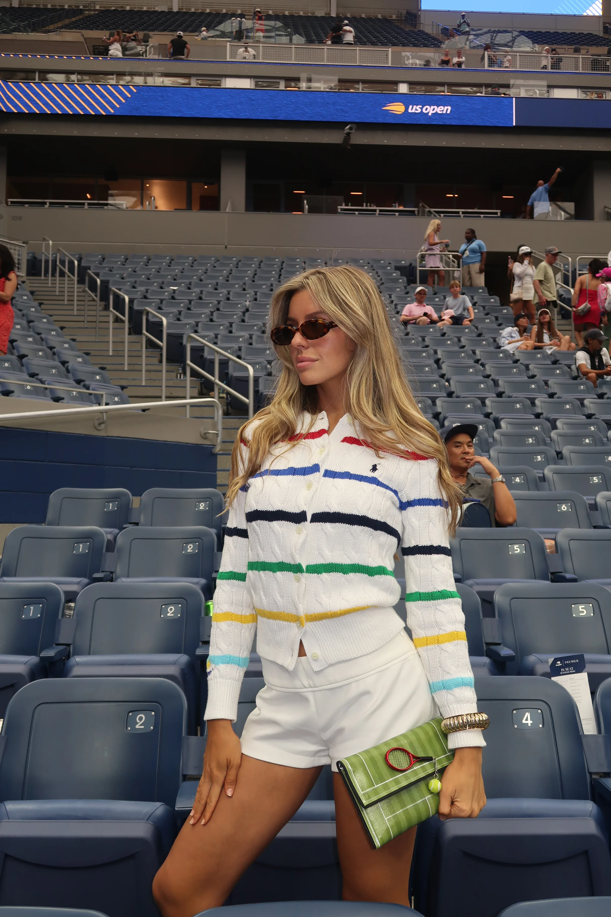 White cable-knit cardigan with colorful stripes, white shorts, brown sunglasses, tennis court clutch.
