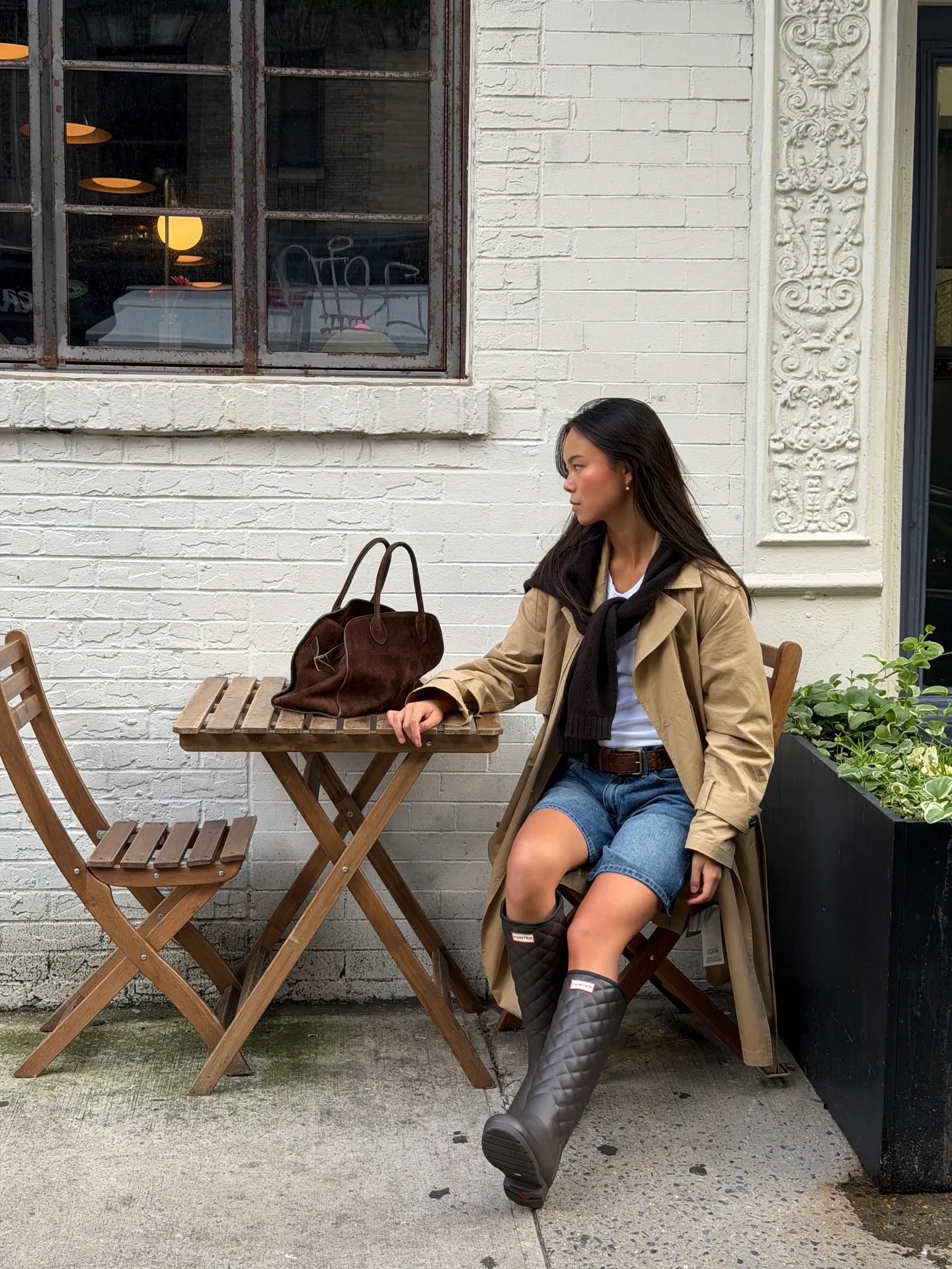 Tan trench coat, denim shorts, black Hunter boots, and brown suede bag at outdoor cafe.
