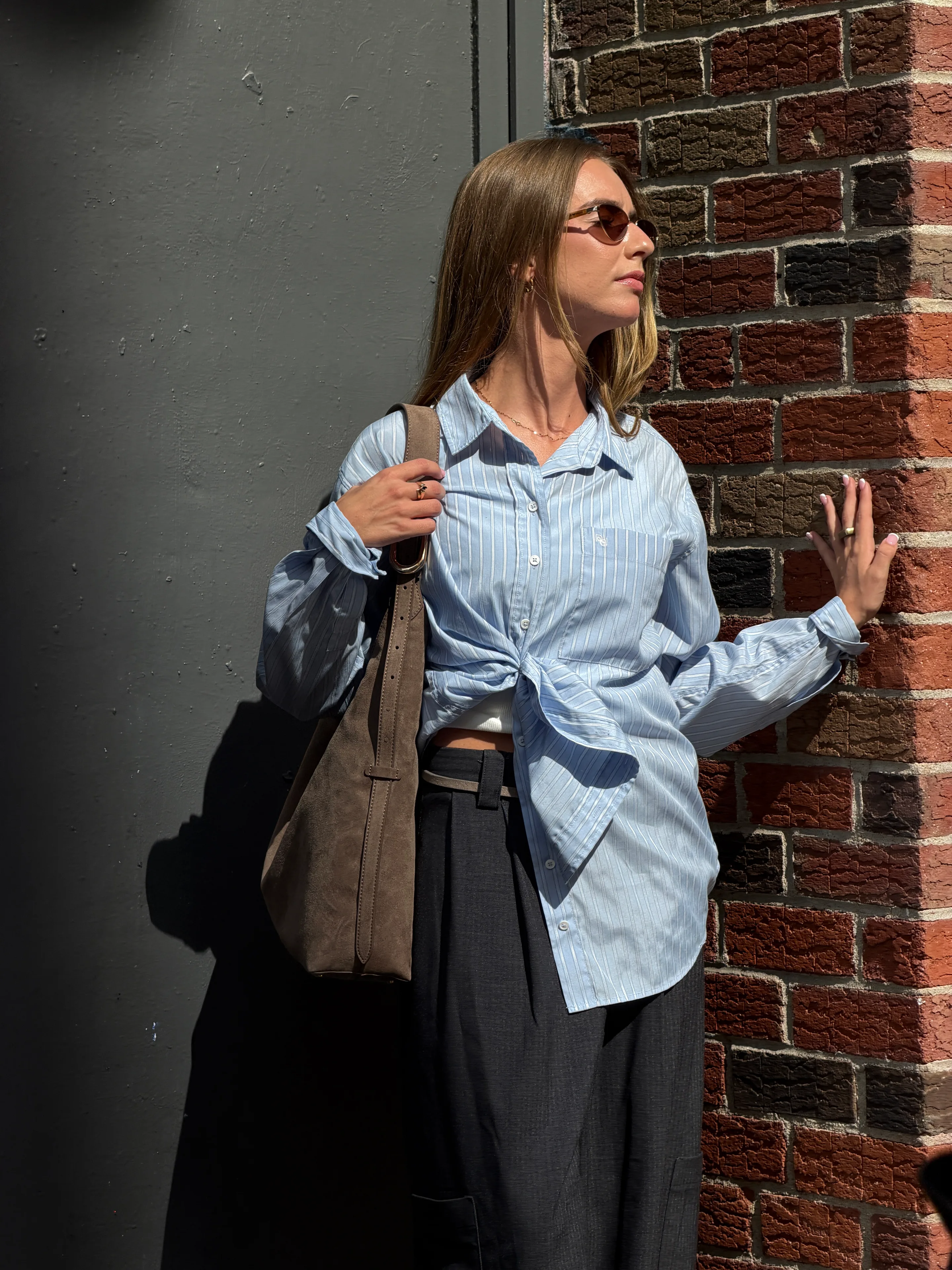 Light blue striped button-down shirt tied at waist, dark wide-leg pants, and brown suede bag.