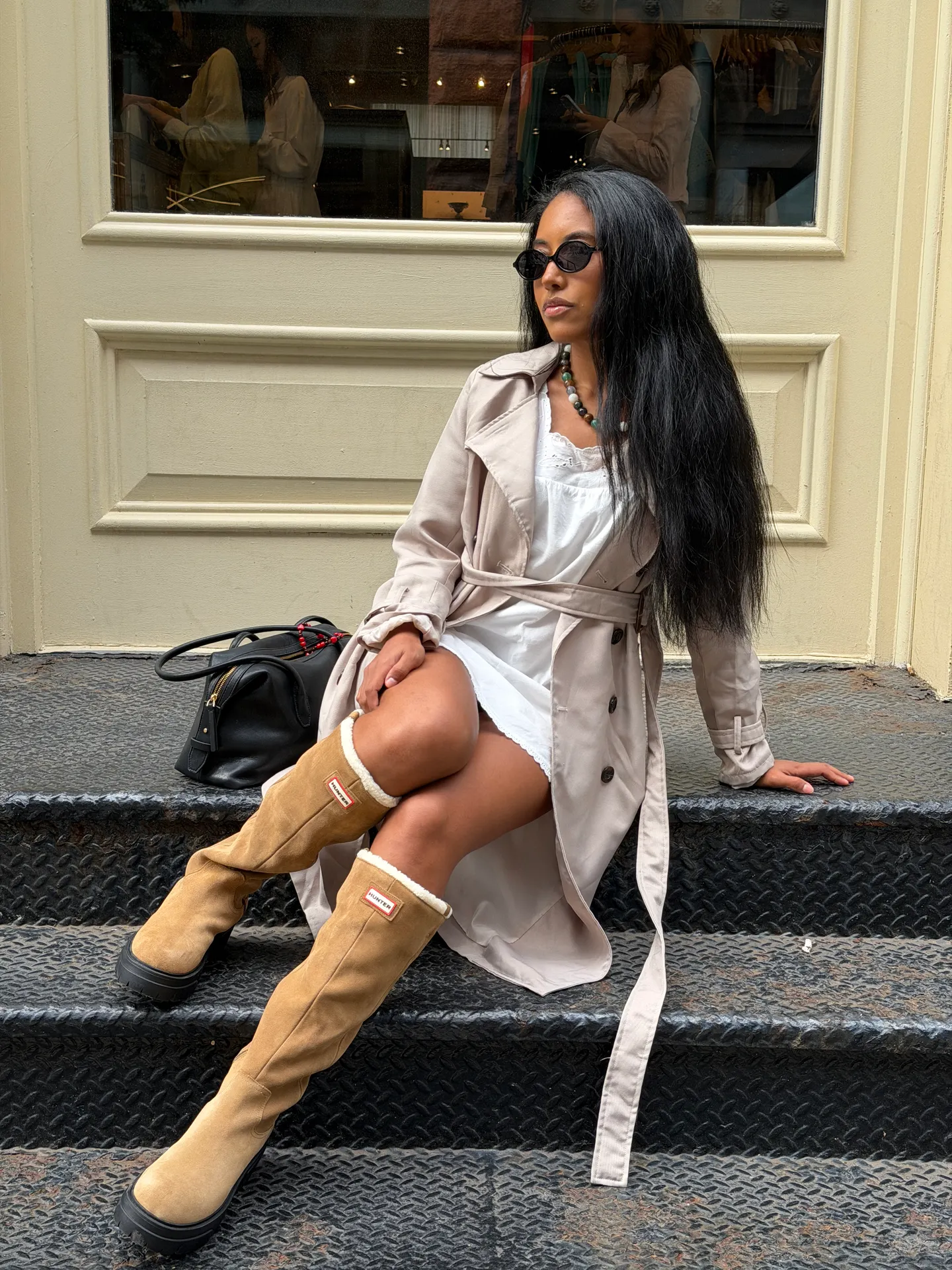 Beige trench coat over a white dress with tan Hunter boots and black sunglasses.