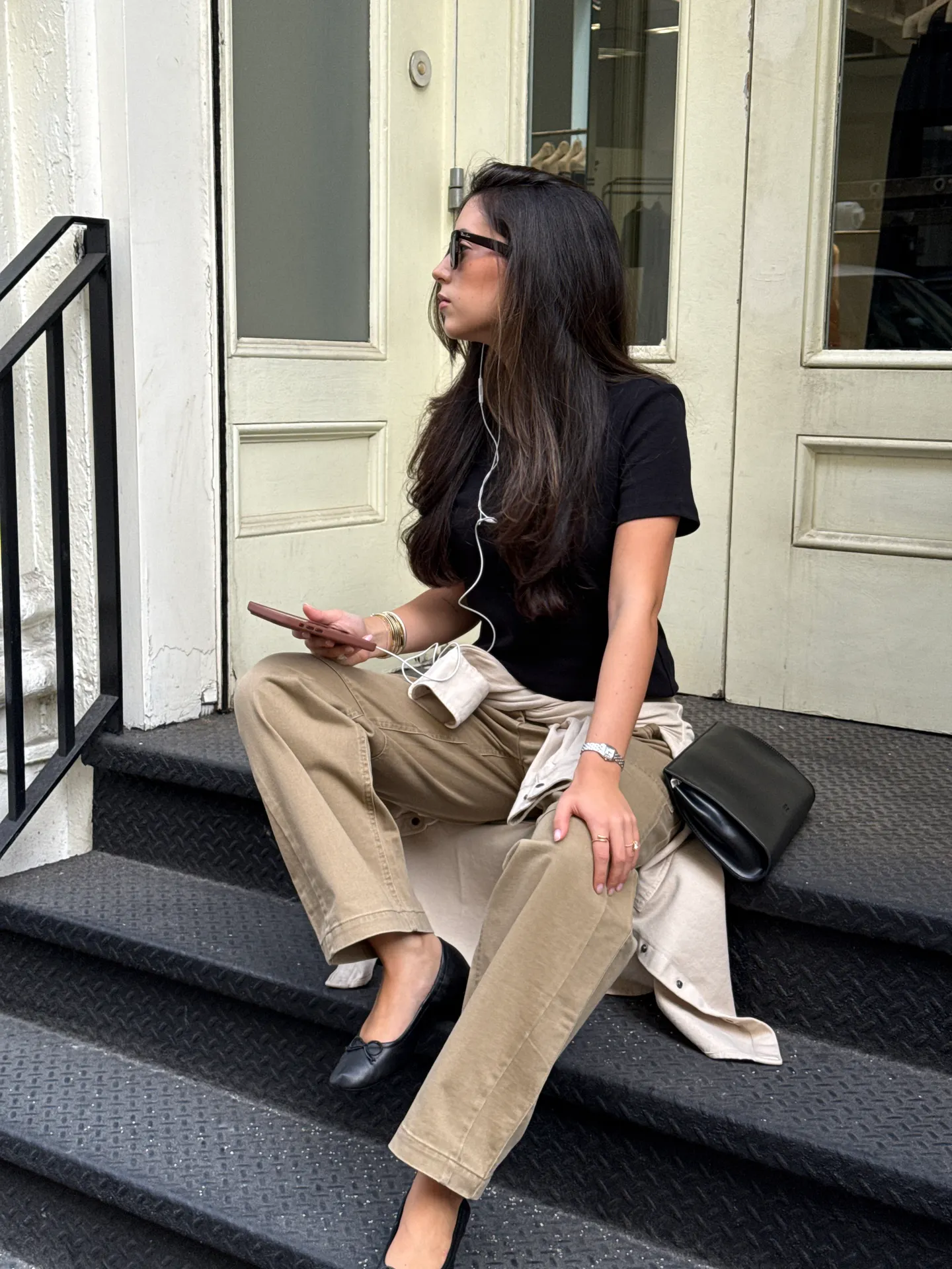 Black t-shirt, khaki pants, and black flats on stairs with sunglasses and a phone.