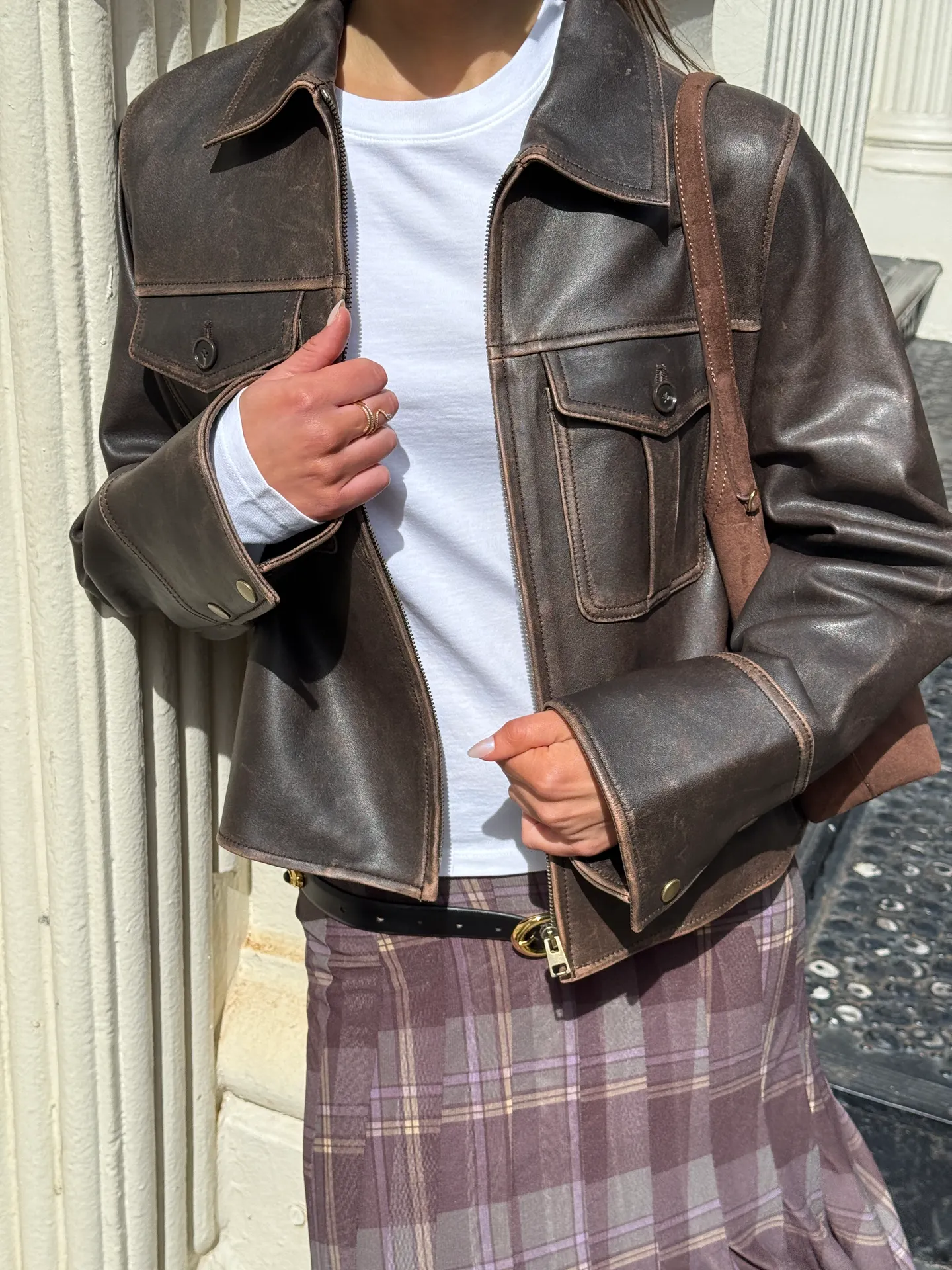 Brown distressed leather jacket over white long-sleeve top and plaid skirt.