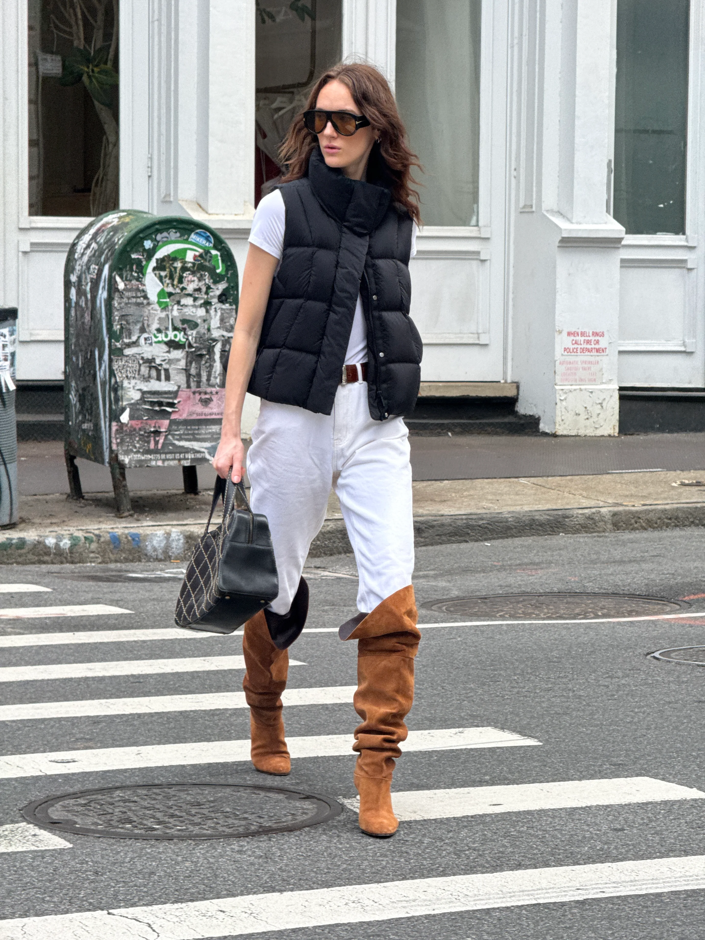 Black puffer vest, white tee, white jeans, brown boots, and sunglasses crossing the street.