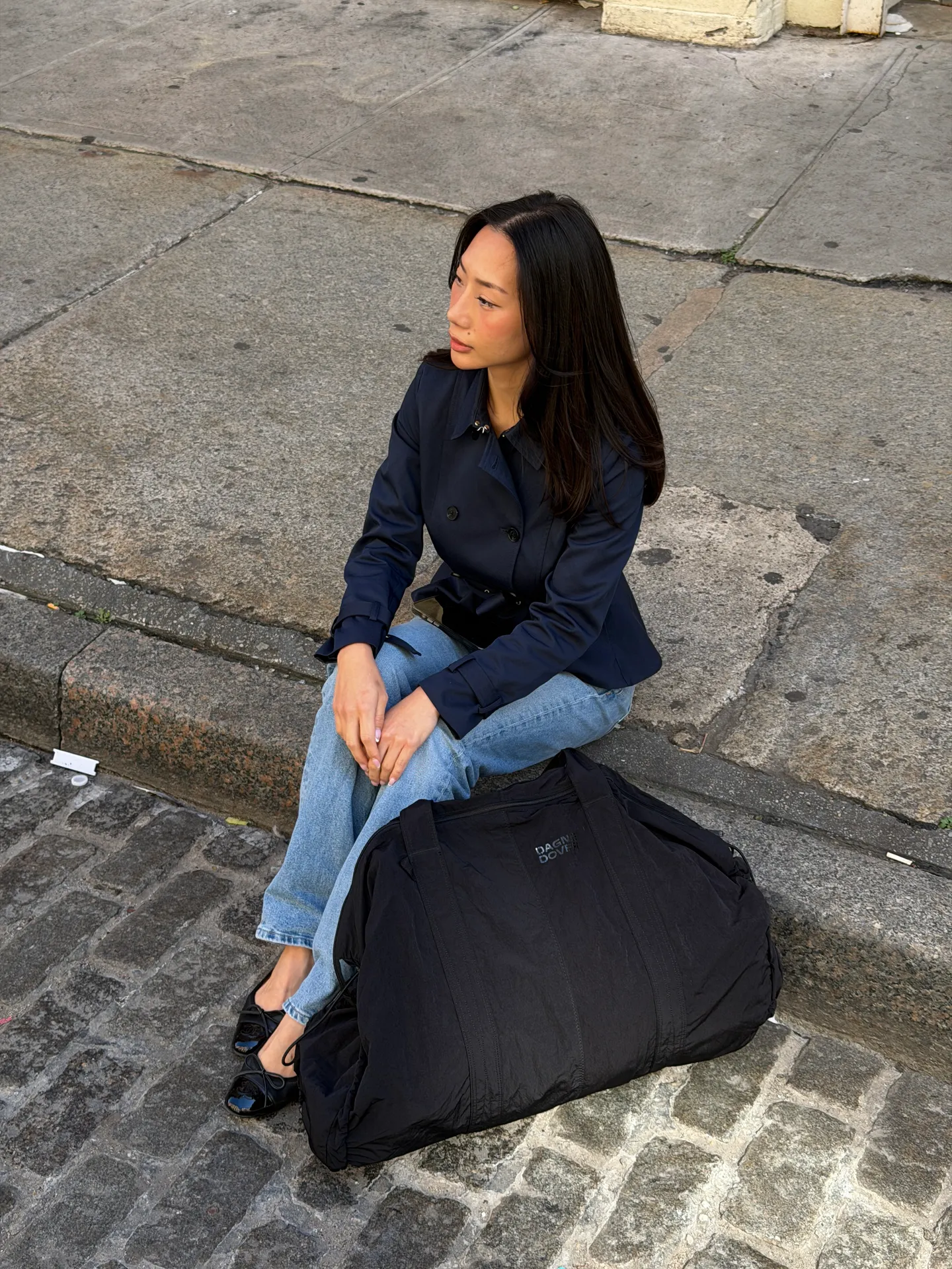 Navy trench coat, light wash jeans, and black ballet flats, sitting on a city curb with a black Dagne Dover bag.