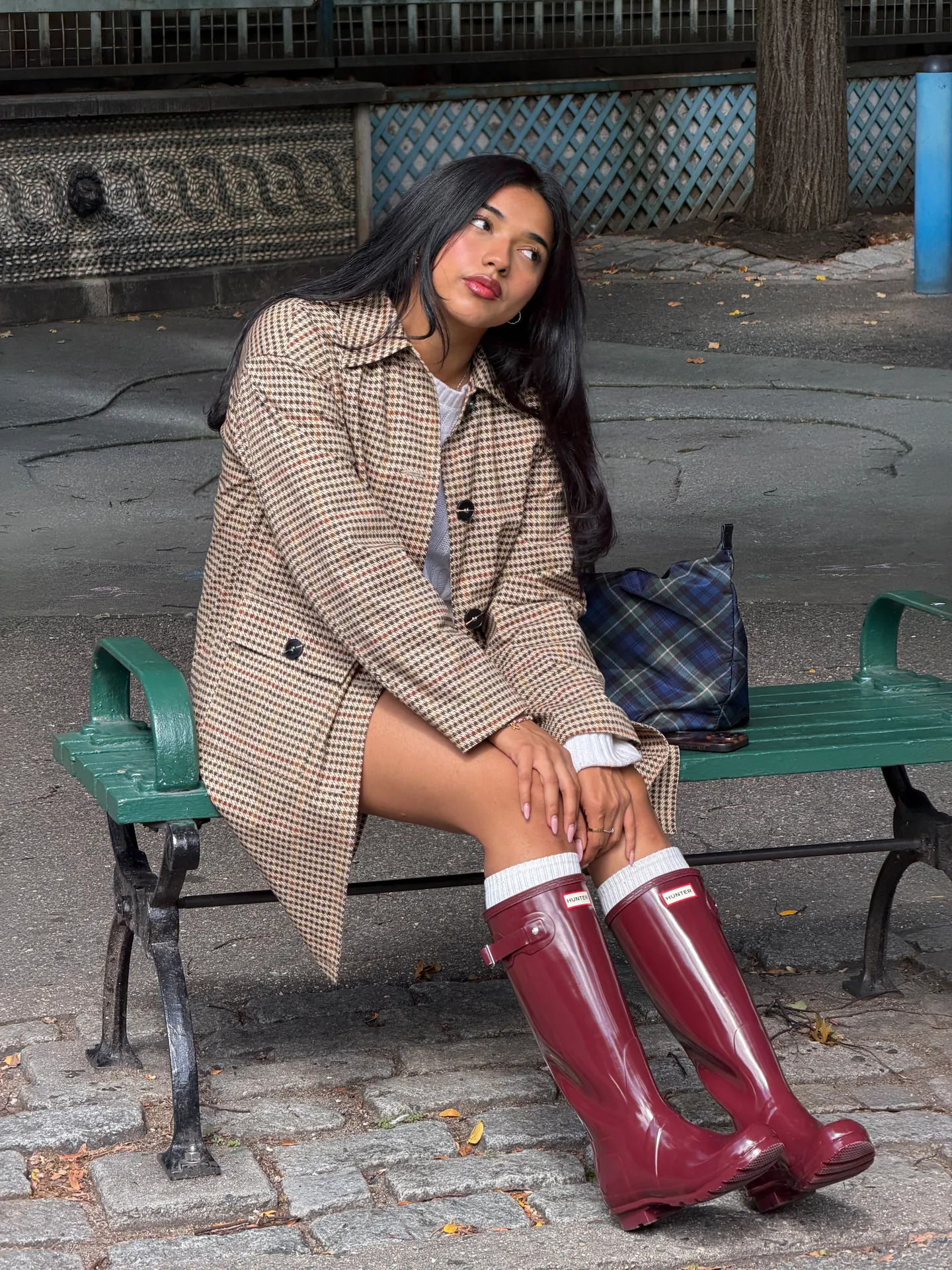 Houndstooth coat, white top, and red Hunter boots with a plaid bag, sitting on a park bench.