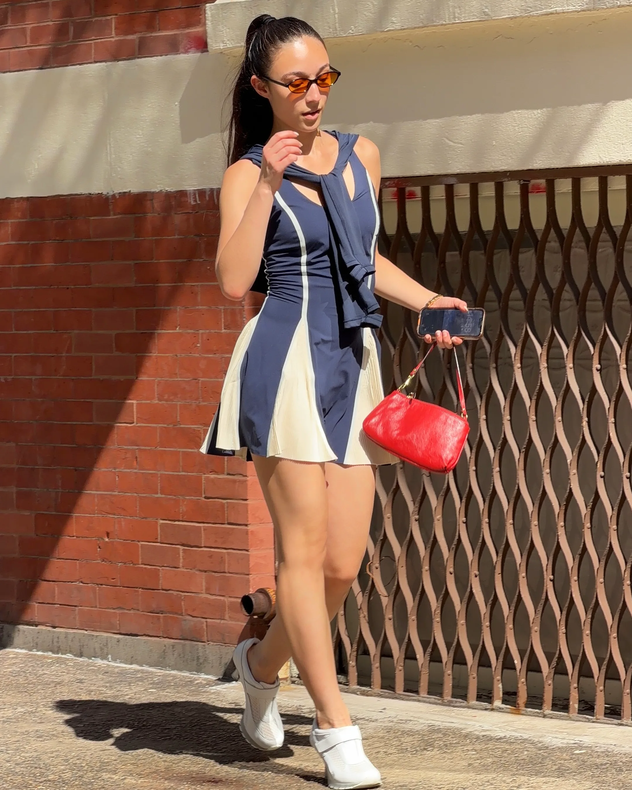 Navy and white mini dress, orange sunglasses, red handbag, and white shoes walking outside.