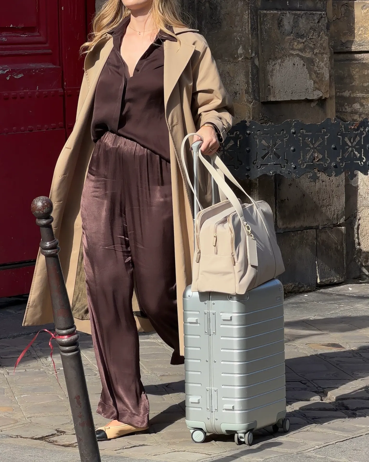 Beige trench coat with brown satin shirt and pants, carrying a light gray suitcase and bag.