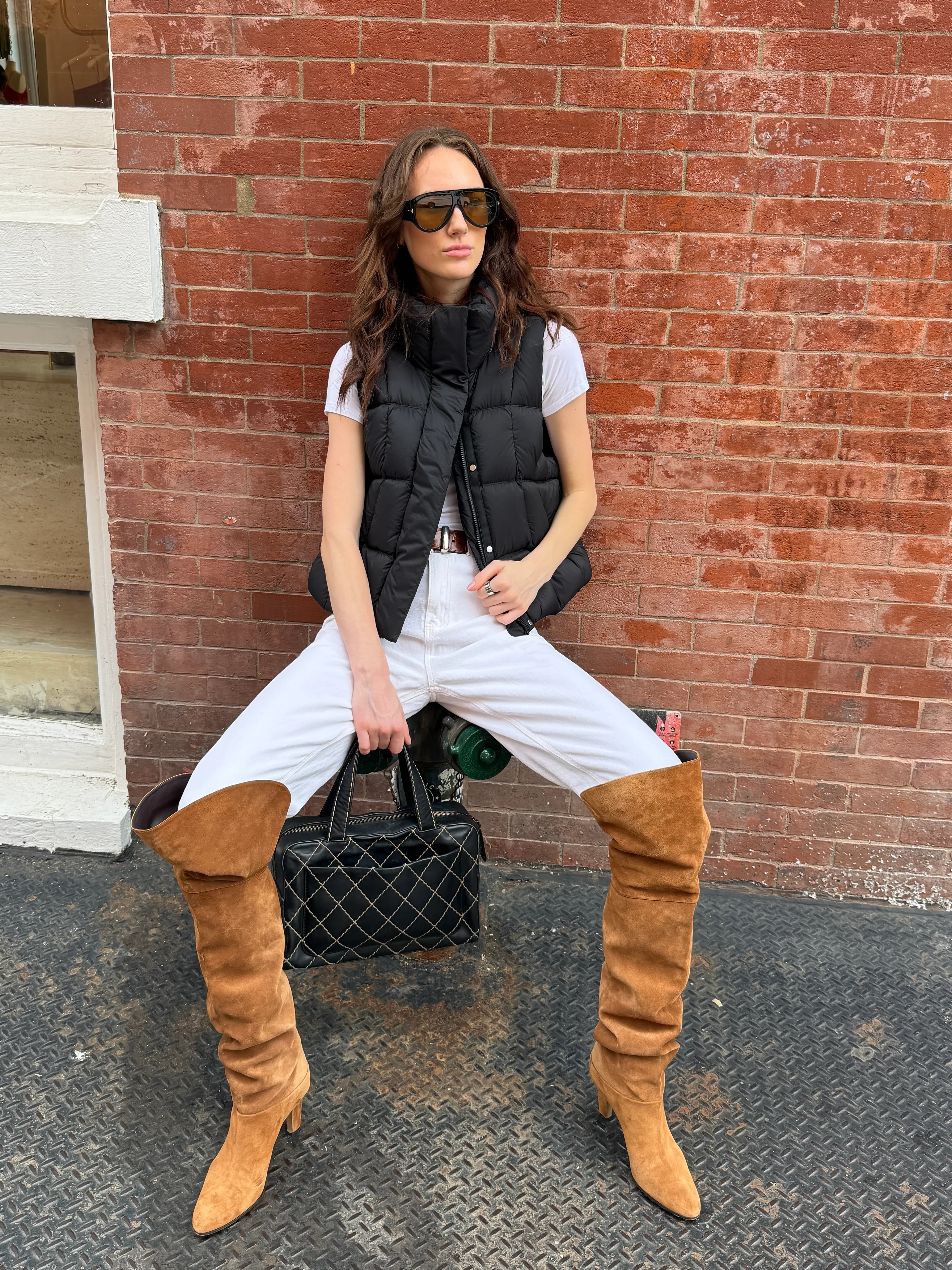 Black puffer vest, white jeans, brown suede over-the-knee boots, and quilted black bag.