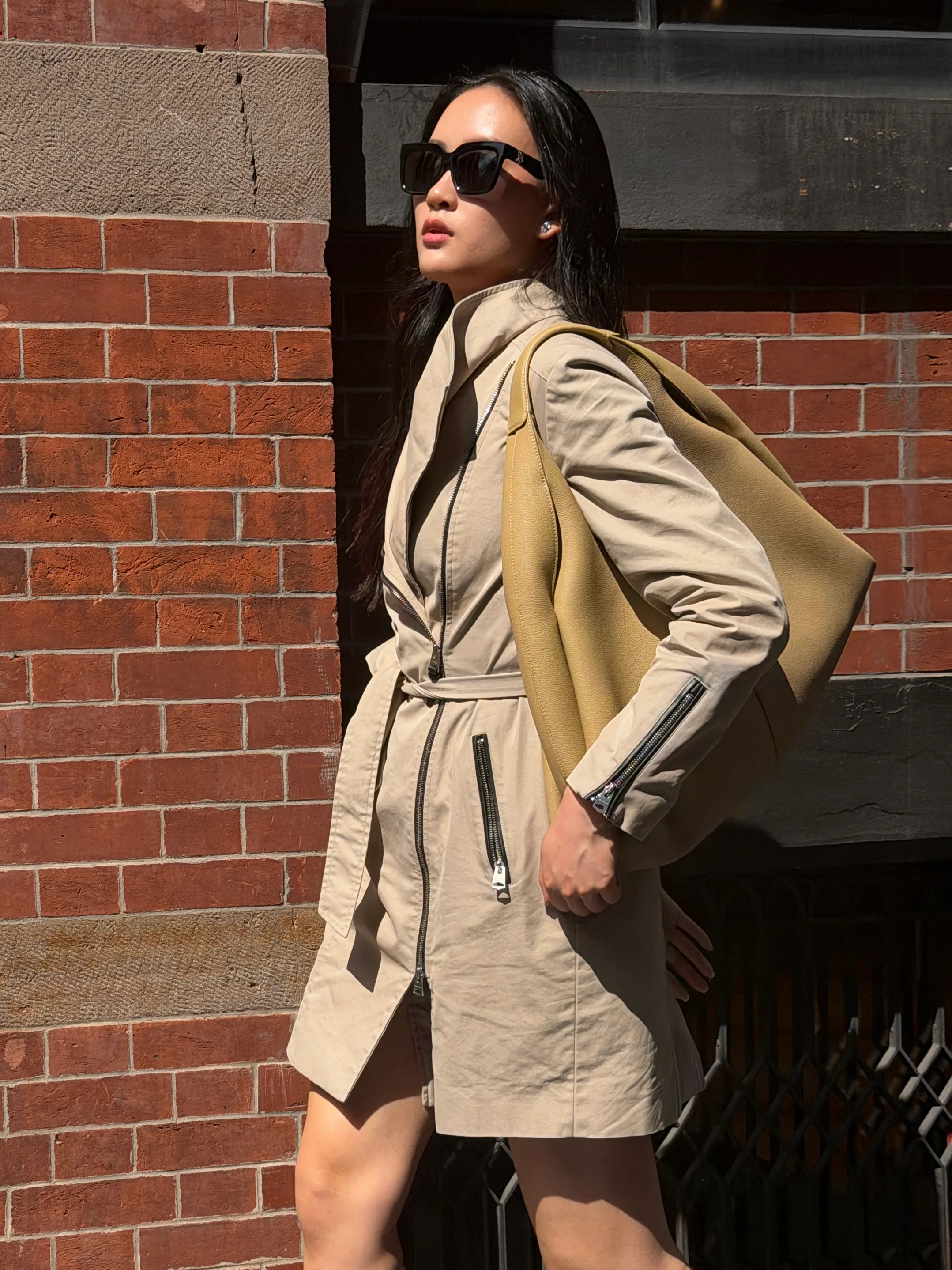 Beige trench coat with black zippers, square sunglasses, and an oversized yellow hobo bag.