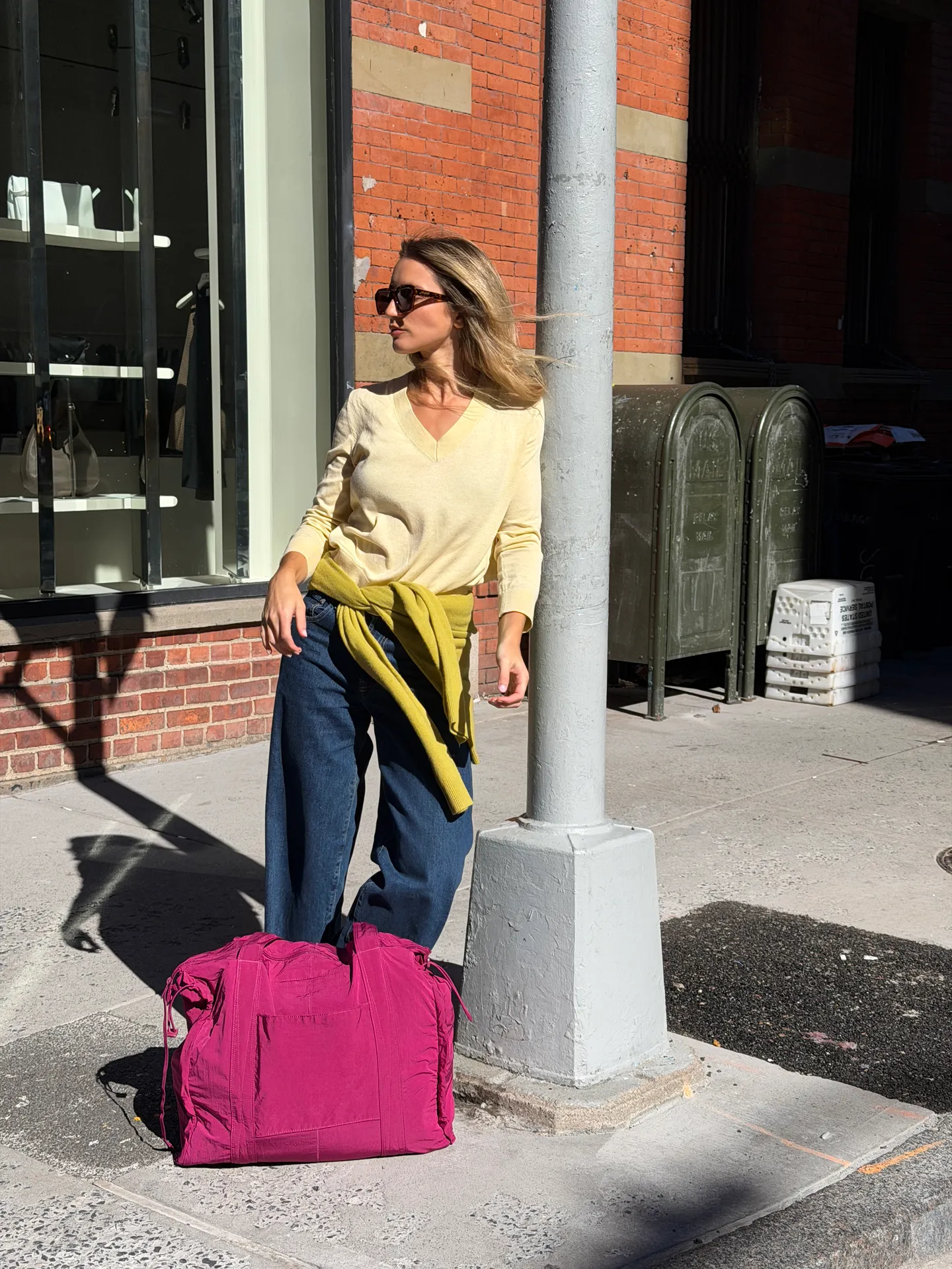 Yellow V-neck sweater, wide-leg jeans, green sweater, and a fuchsia duffel bag on a city street.