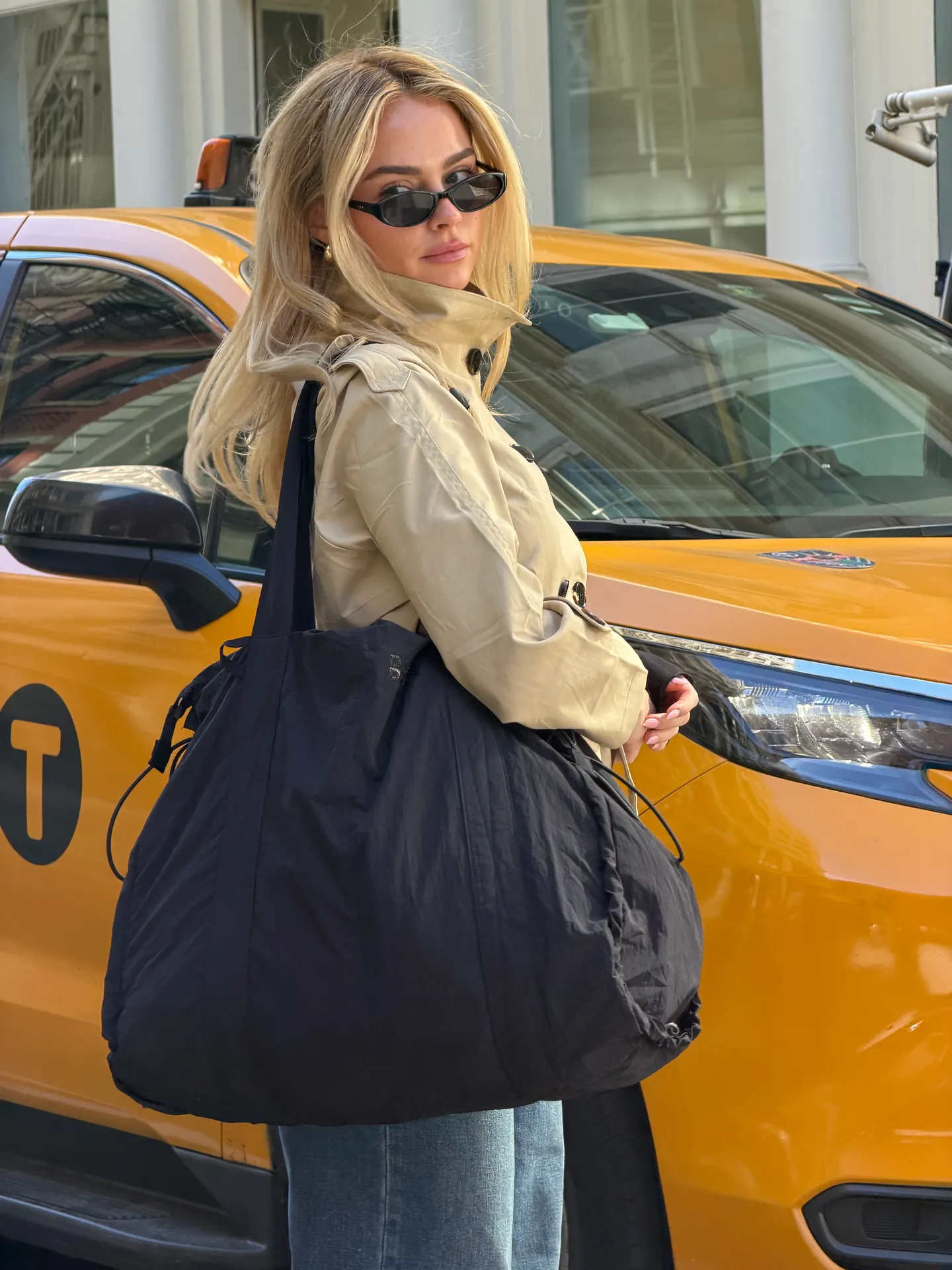 Tan trench coat, black cat-eye sunglasses, and black oversized tote bag by a yellow taxi.