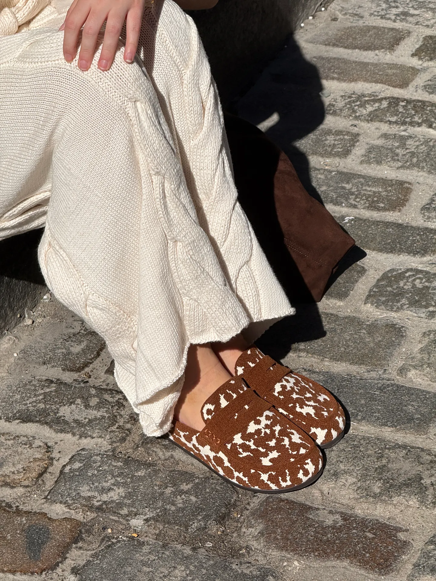 Cable-knit cream sweater pants with brown and white cow print slippers on cobblestone.