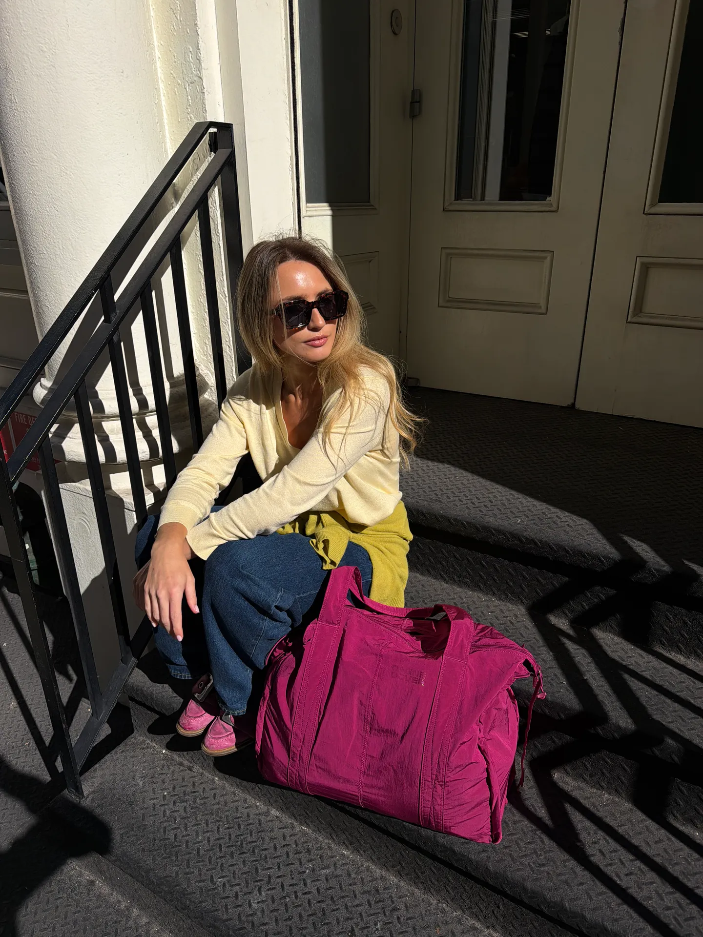 Blonde woman in yellow cardigan, blue jeans, and pink shoes on steps with a fuchsia bag.