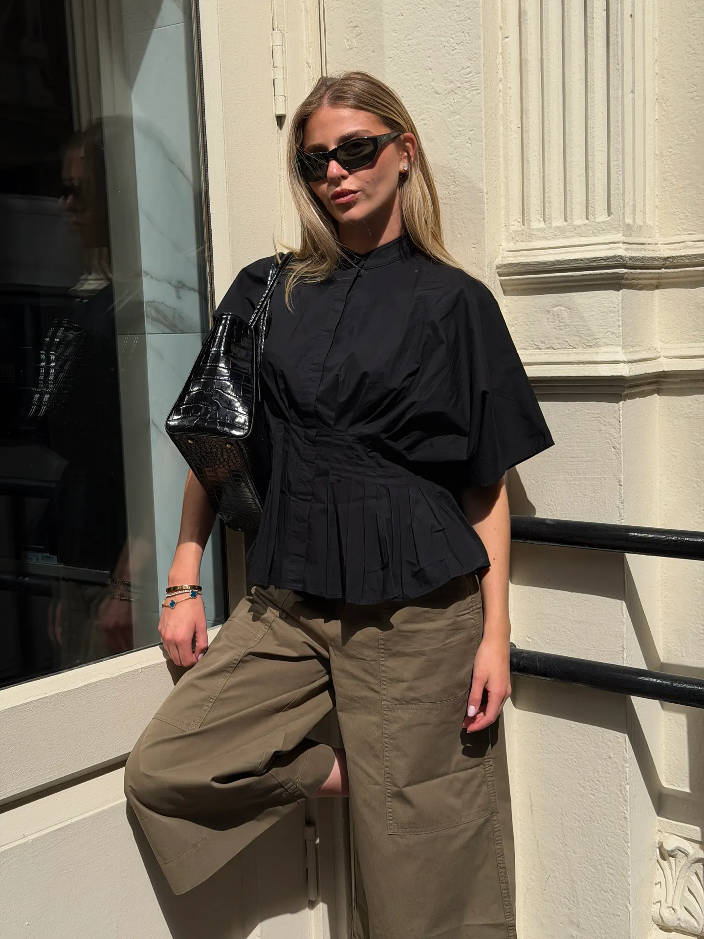 Black pleated peplum top, olive green wide-leg pants, black croc-embossed bag, and sunglasses.