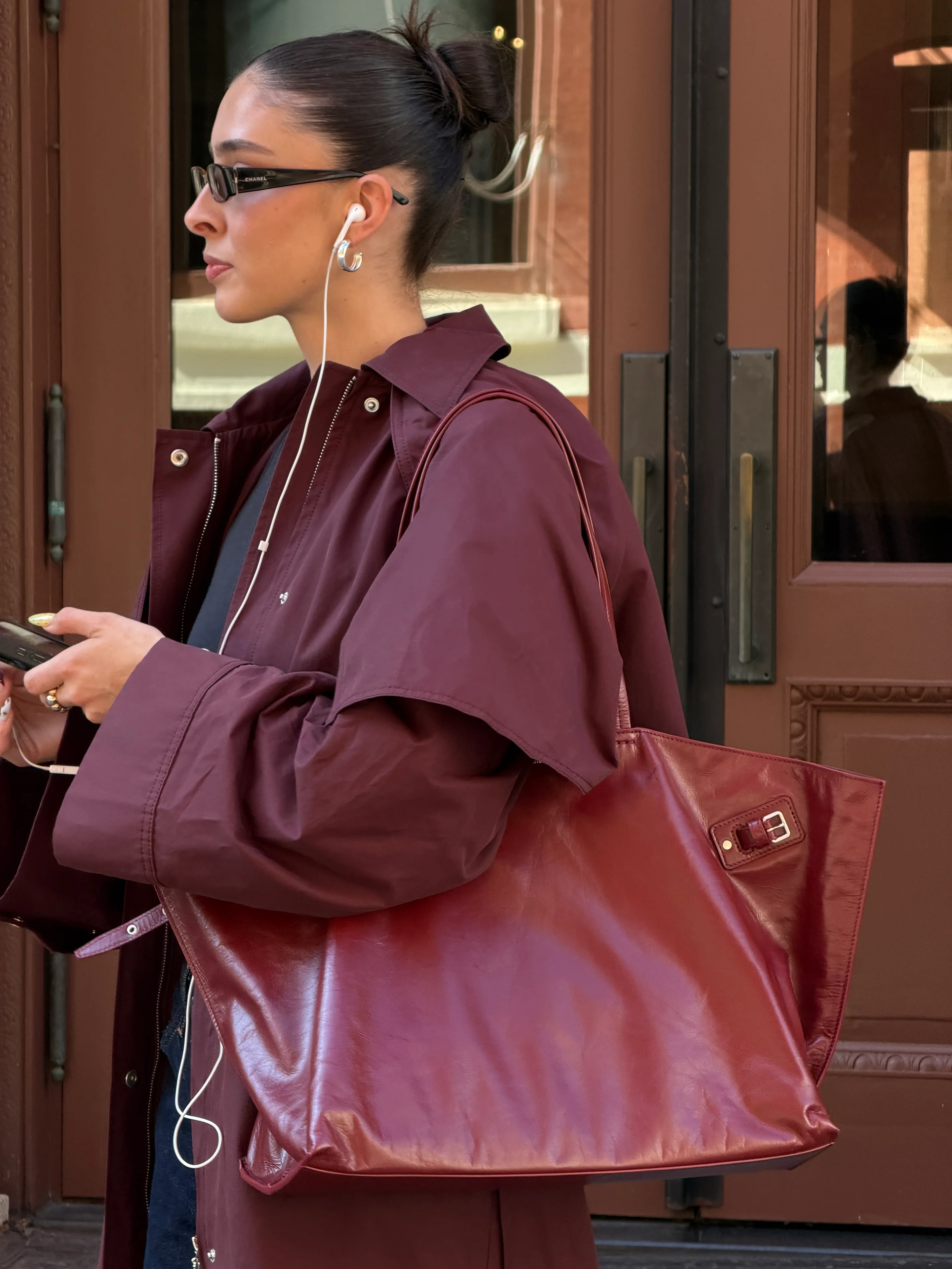 Burgundy coat and shiny tote bag, black sunglasses, hoop earrings, and wired earbuds.