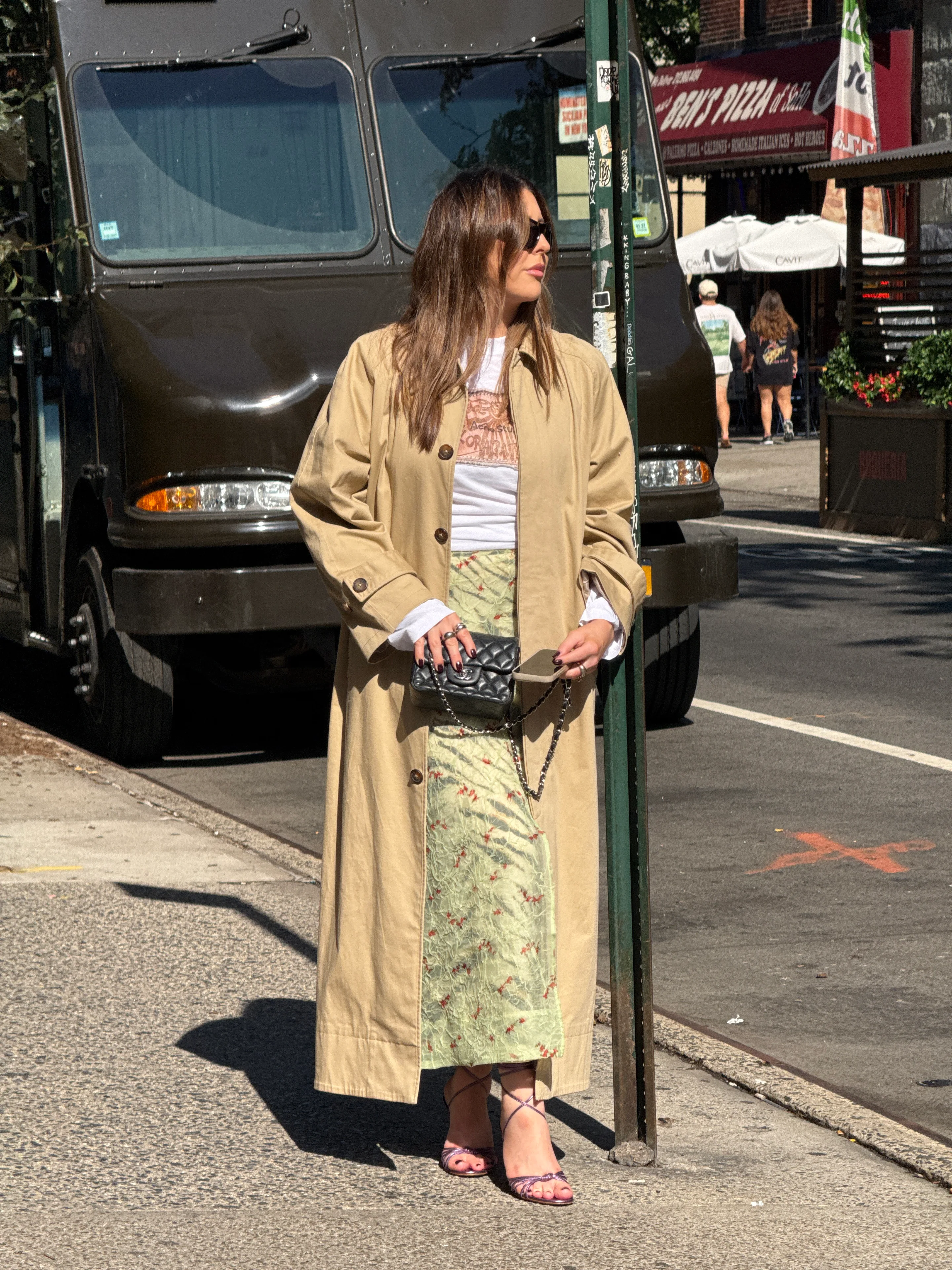 Khaki trench coat over a white tee and green floral skirt, with a black quilted bag.