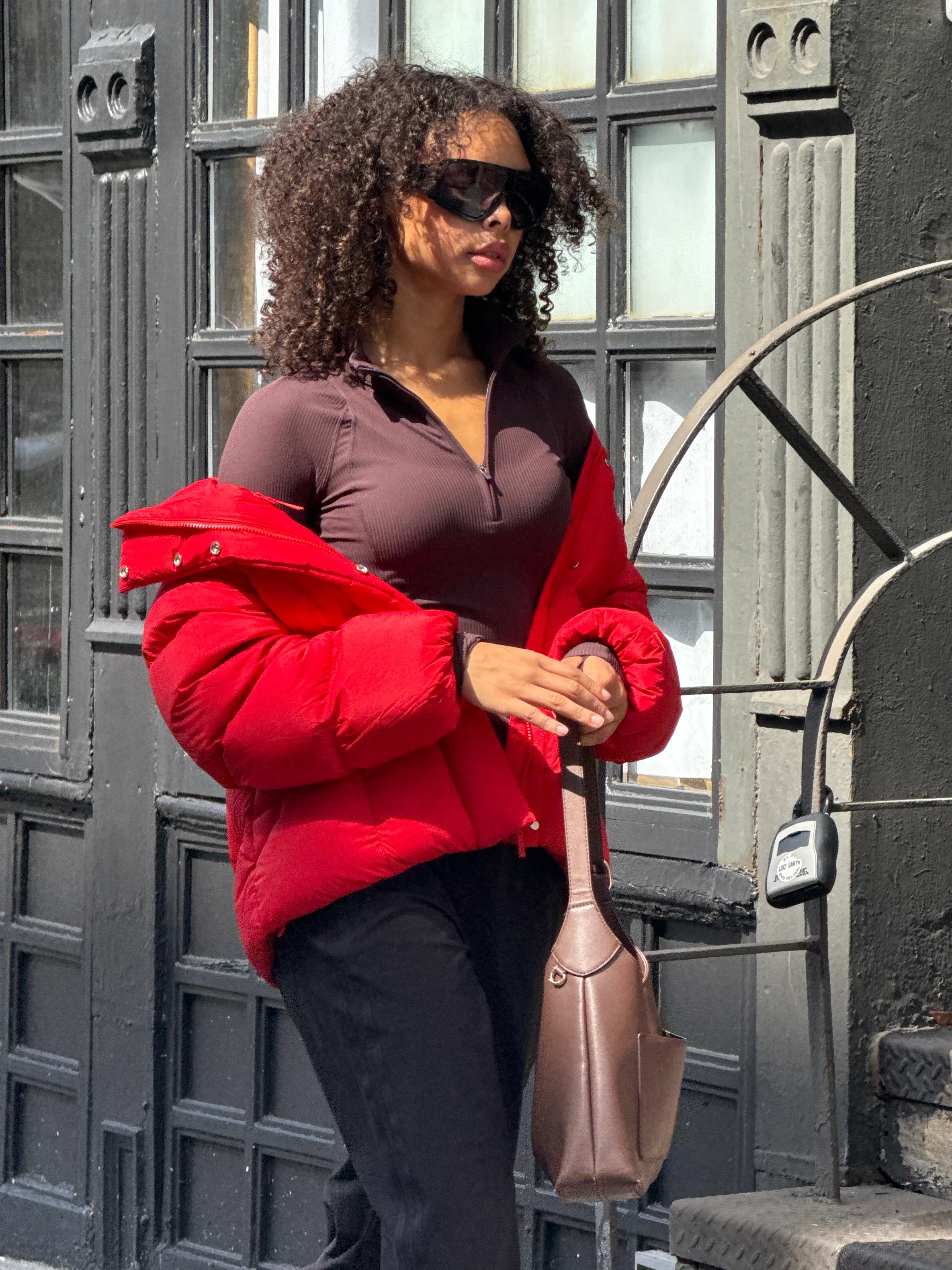Red puffer jacket with brown ribbed top, black pants, brown handbag, and sunglasses.