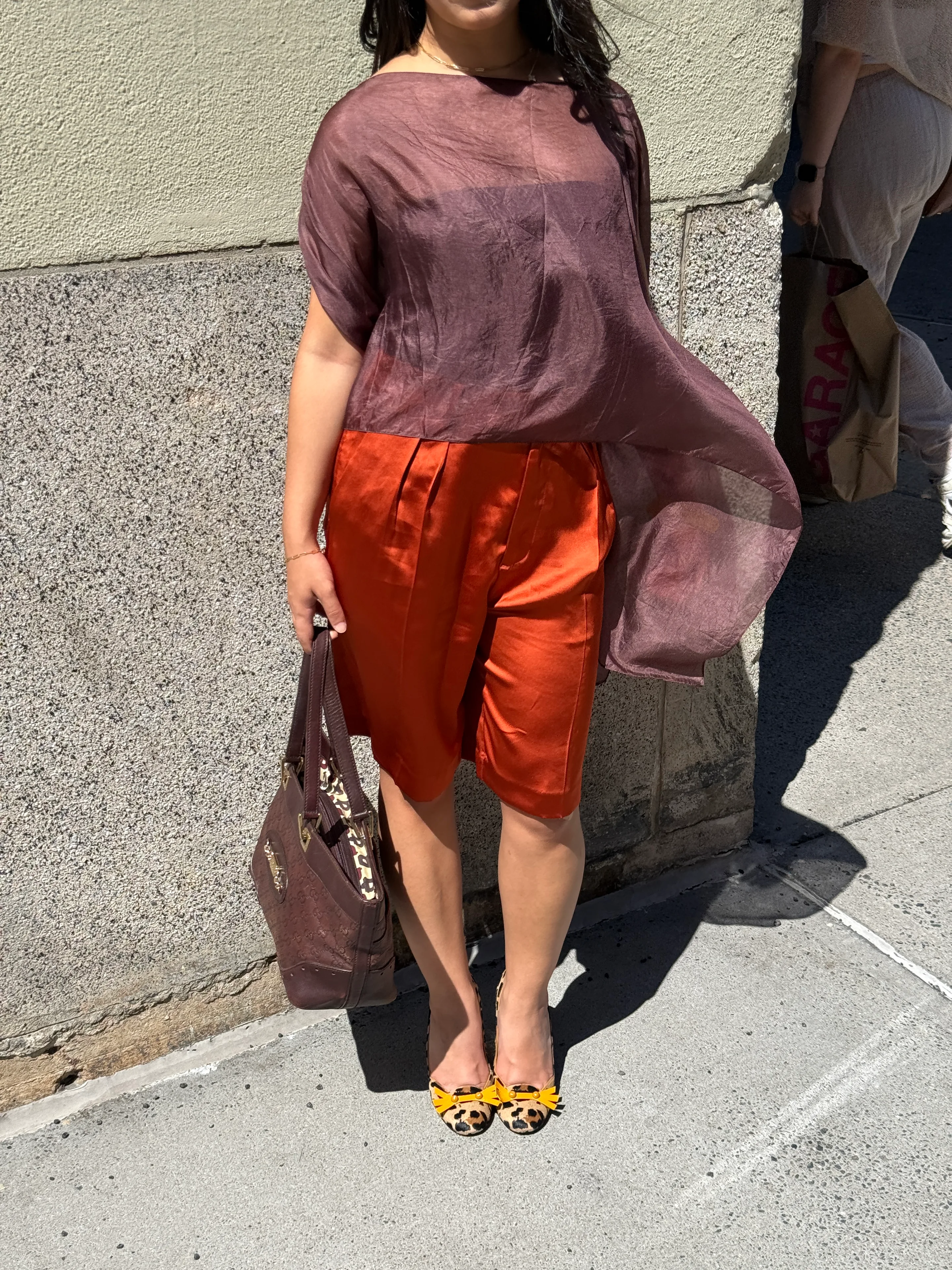 Sheer aubergine top, orange satin shorts, leopard print flats with yellow bows, and a brown bag.