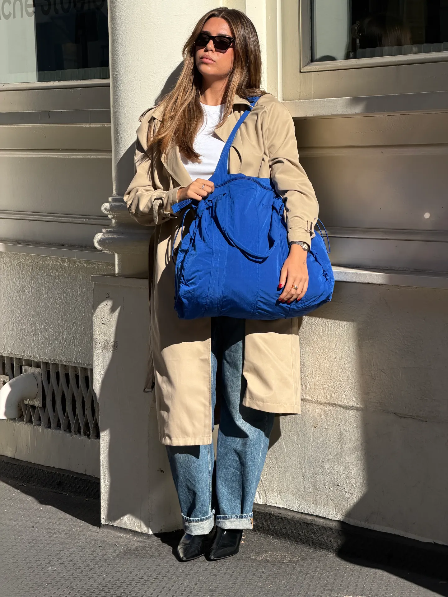 Beige trench coat with straight-leg jeans, black boots, and a royal blue tote bag.