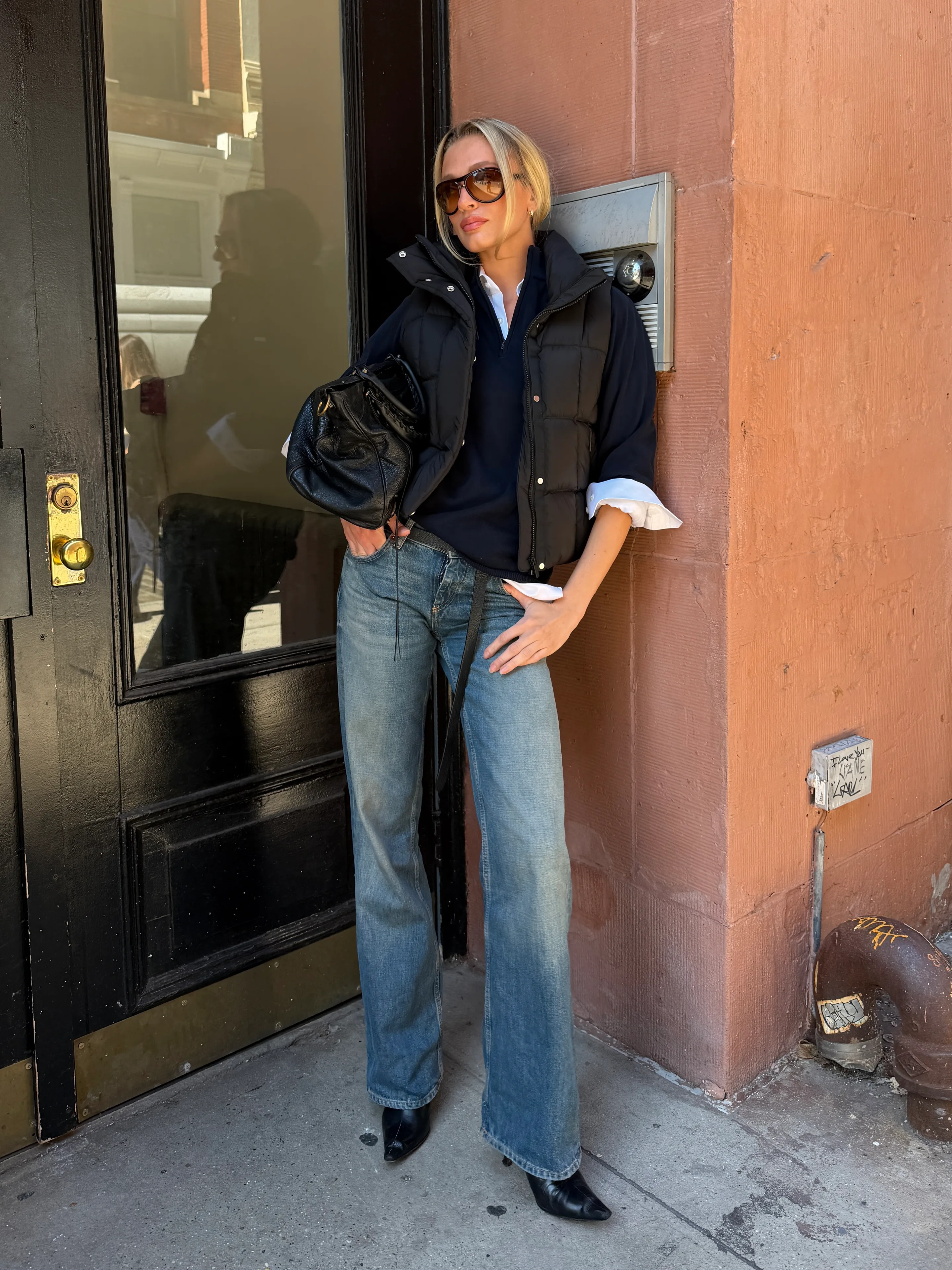 Black puffer vest over blue sweater and white shirt, blue jeans, black boots.
