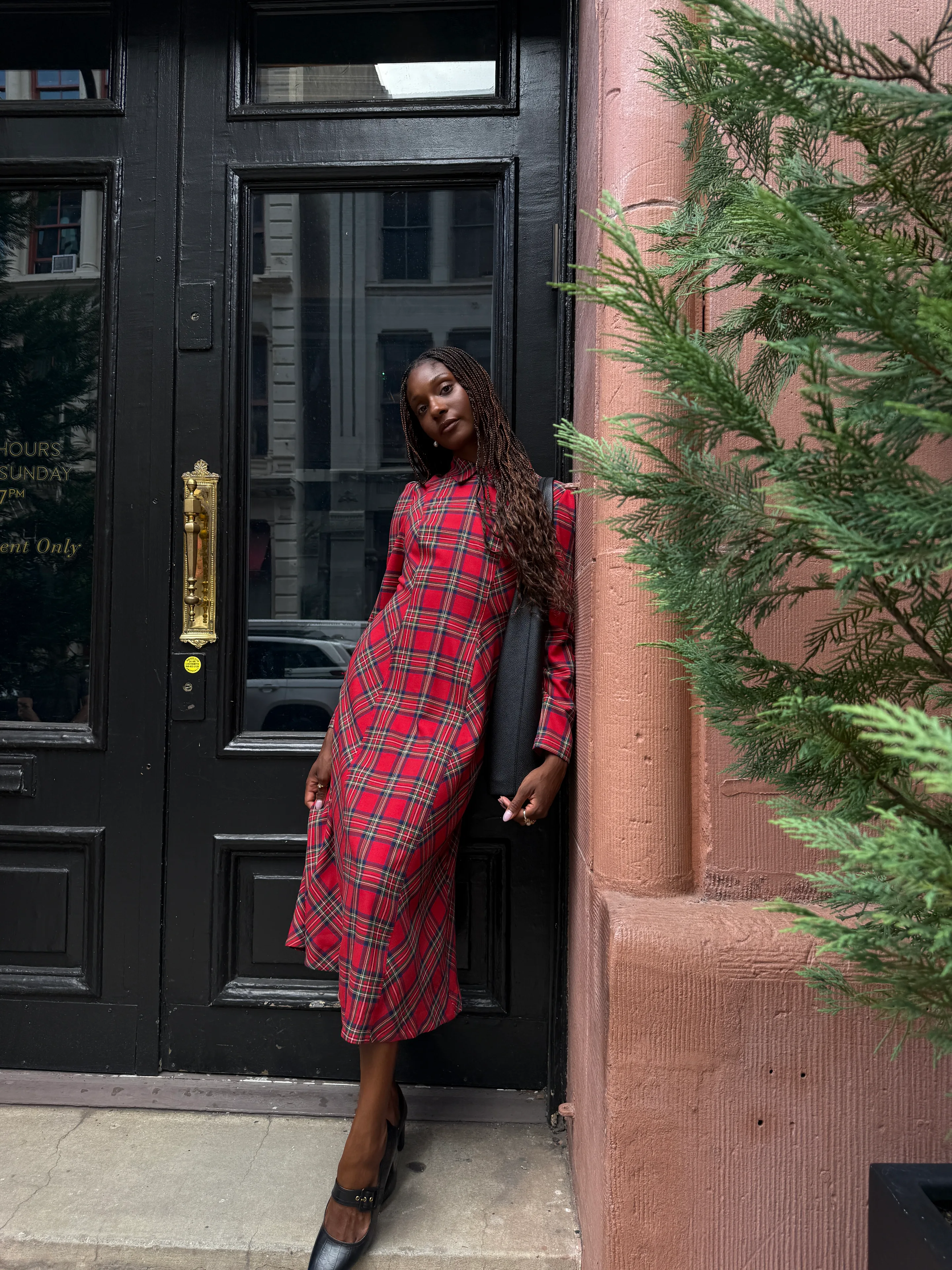 Red plaid collared midi dress with black mary jane shoes, leaning against a black door.