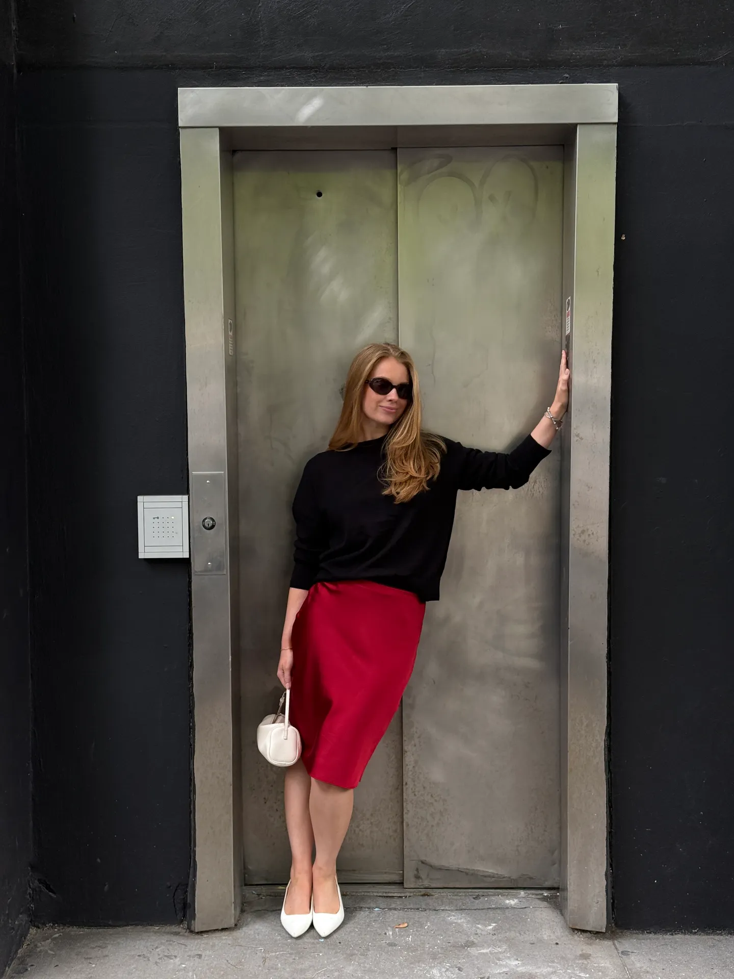 Black long-sleeve top with red midi skirt, white pumps, and white handbag by an elevator.