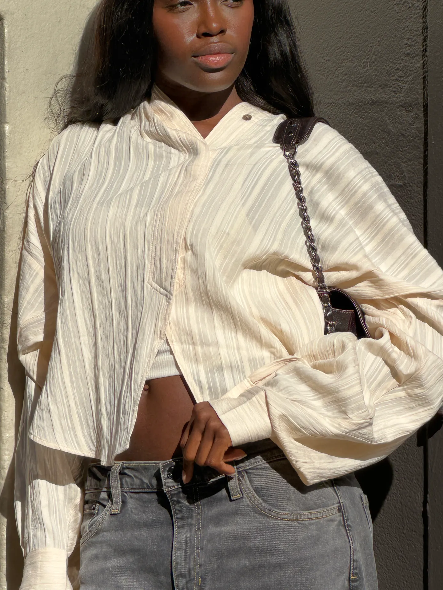 Cream pleated button-up shirt, gray washed jeans, and a dark chain-strap handbag.