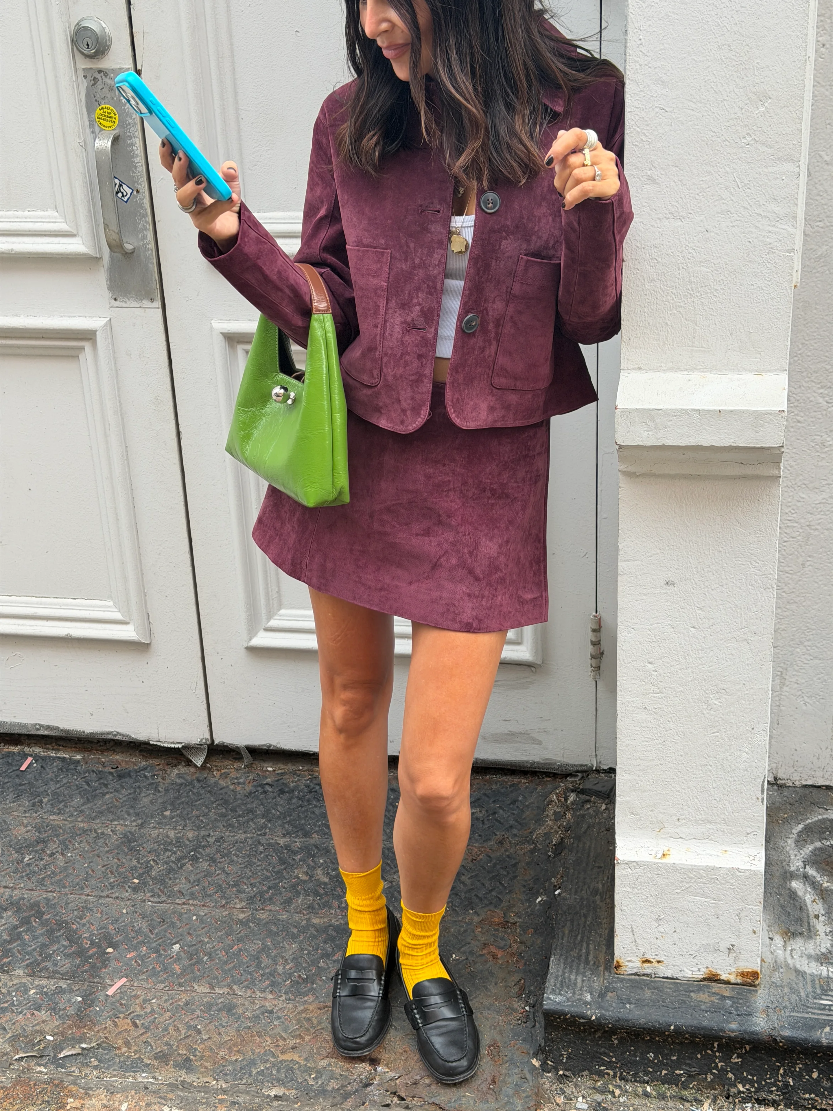 Plum suede cropped jacket and mini skirt, with a bright green handbag and blue phone.