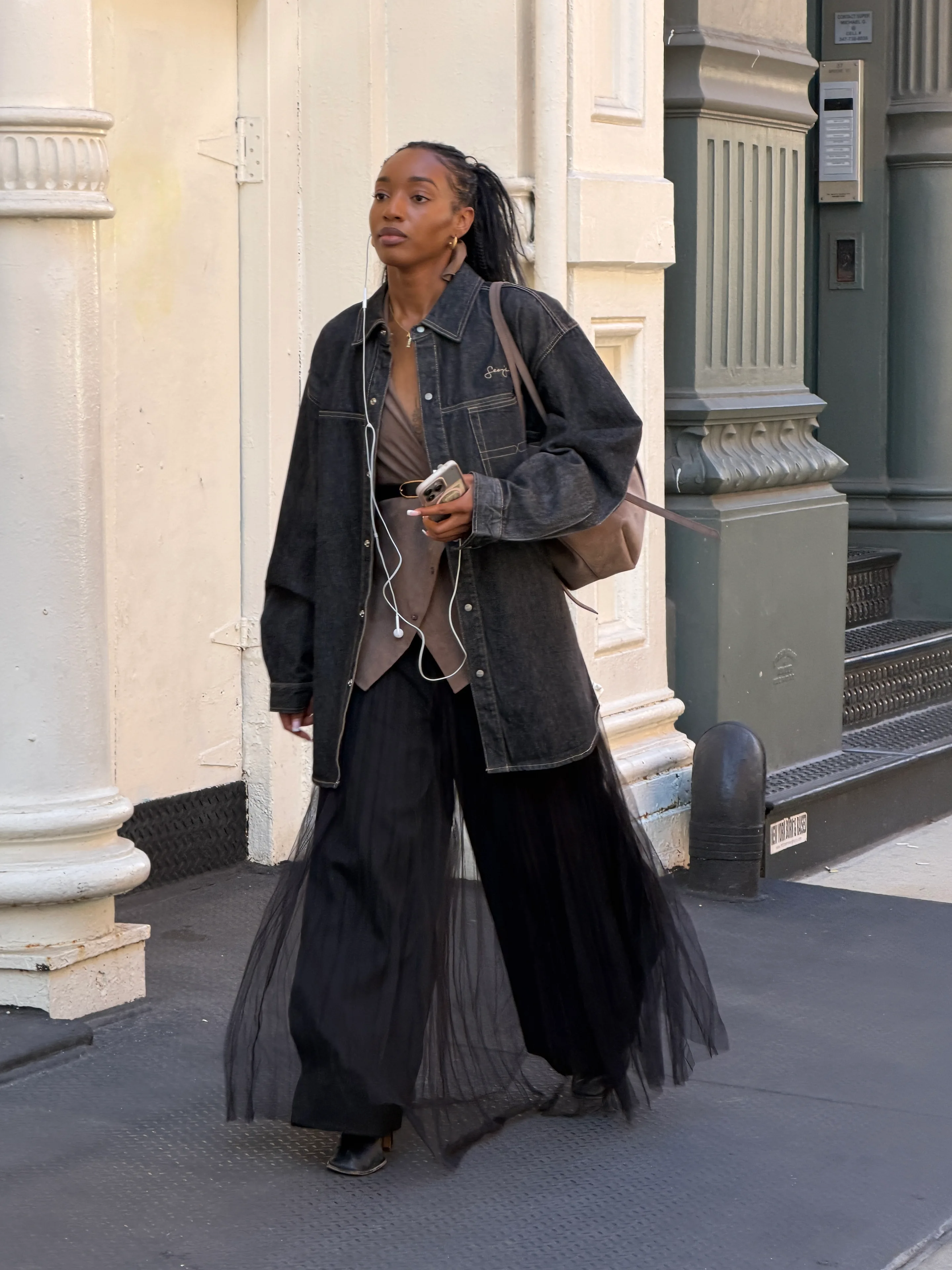 Washed black denim jacket, taupe top, and flowing black sheer-overlay pants on a city street.