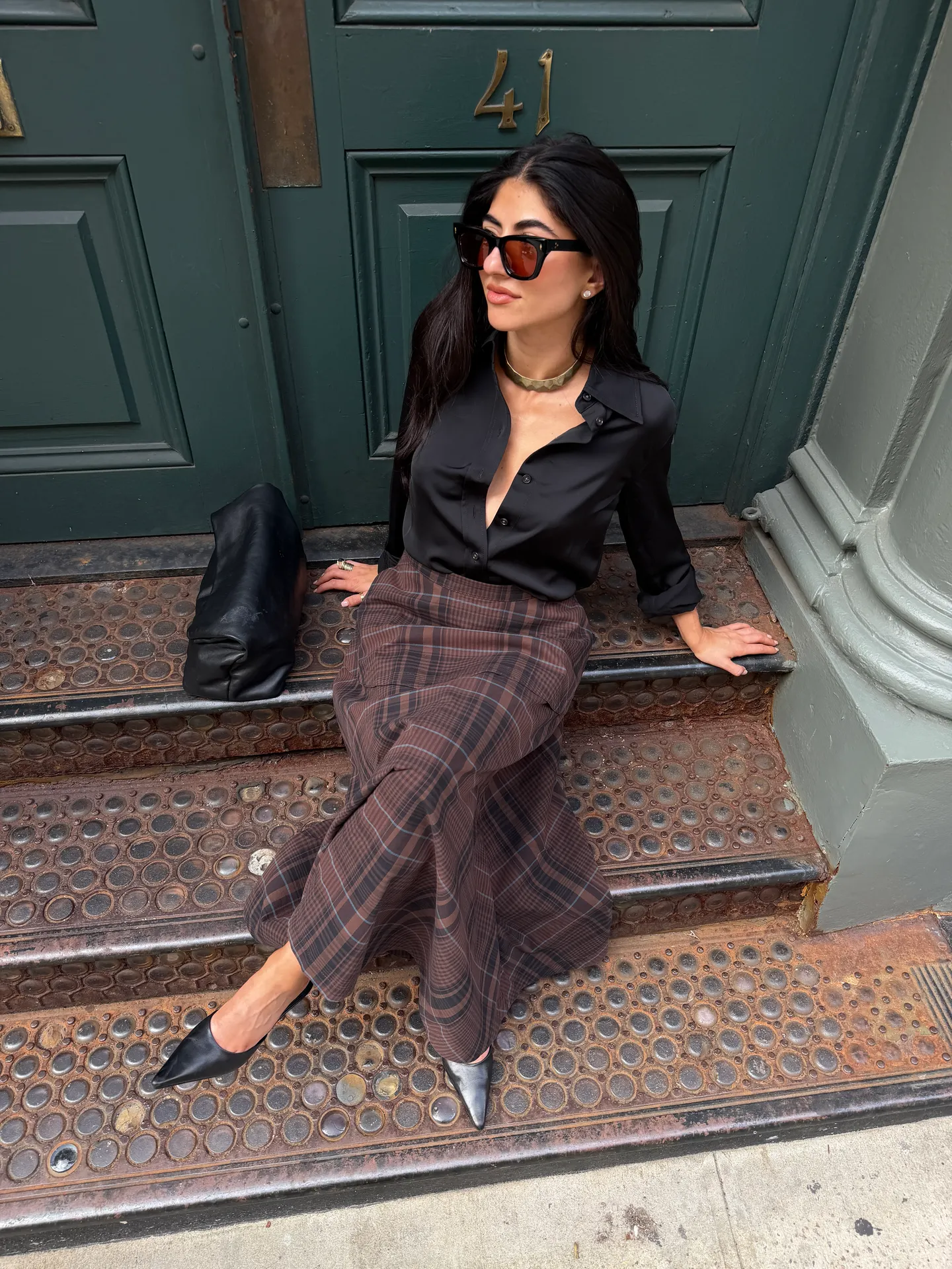 Black satin shirt, brown plaid midi skirt, and black pointed-toe heels sitting on steps.