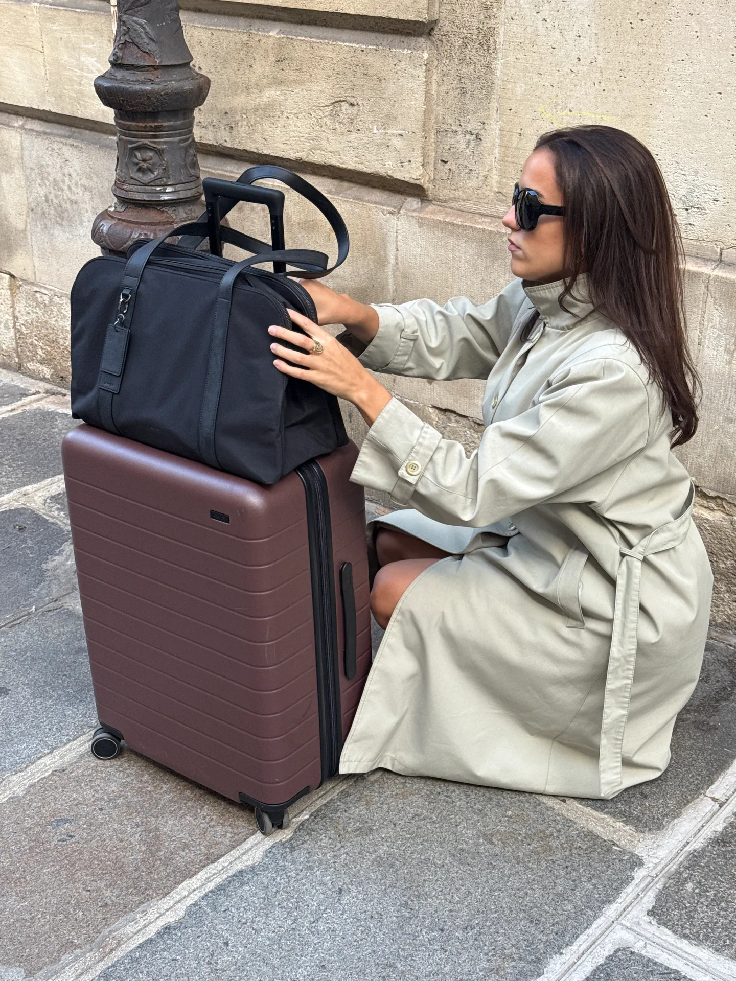 Beige trench coat with black sunglasses, brown suitcase, and black duffel bag on stone pavement.