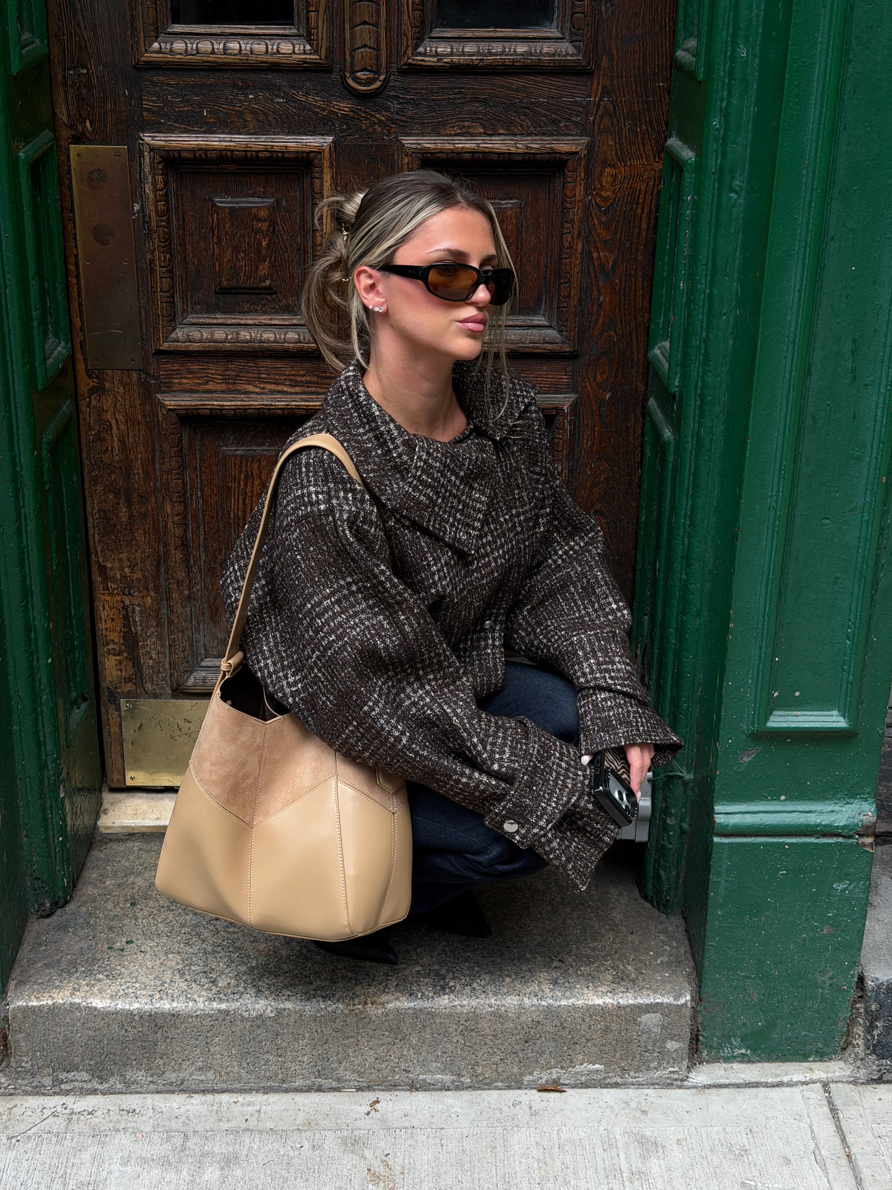 Brown tweed jacket, dark jeans, and a beige shoulder bag, squatting by a wooden door.