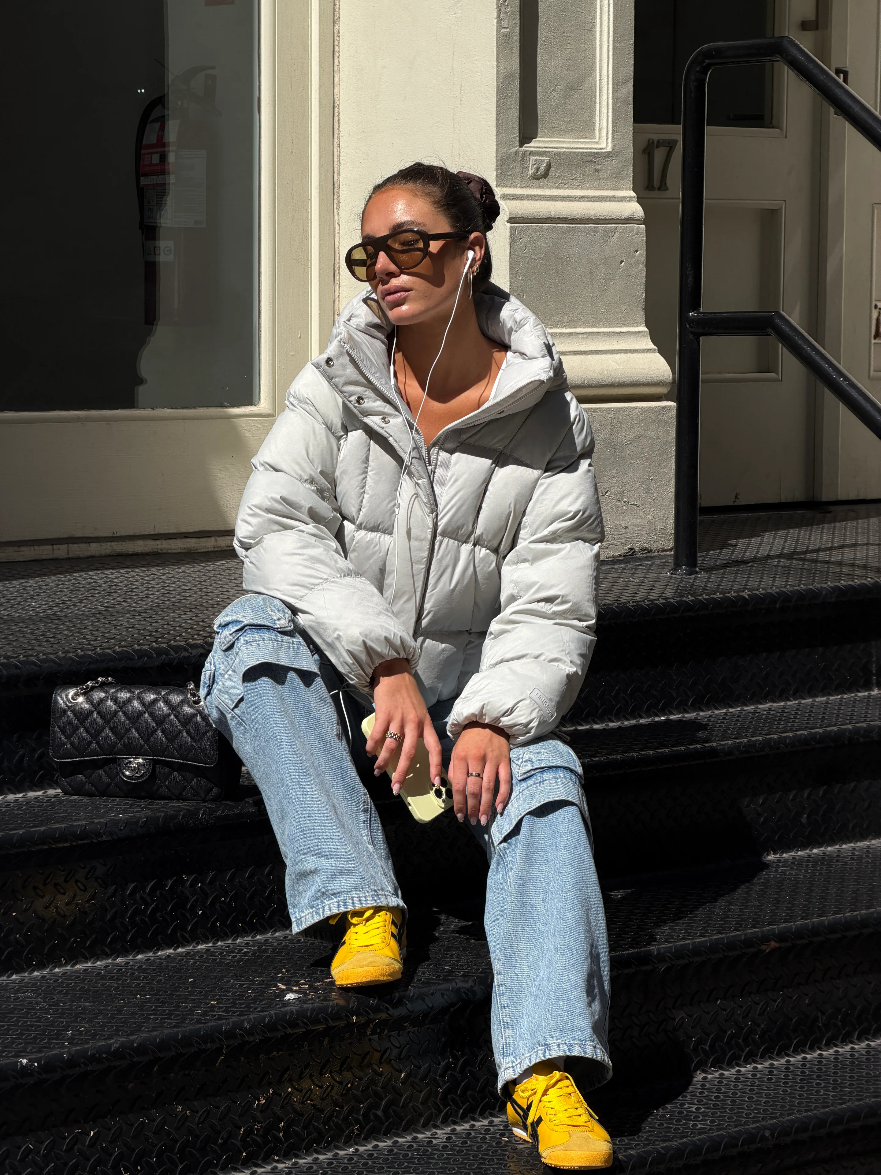 Light gray puffer jacket, light wash jeans, yellow sneakers, and black quilted bag.