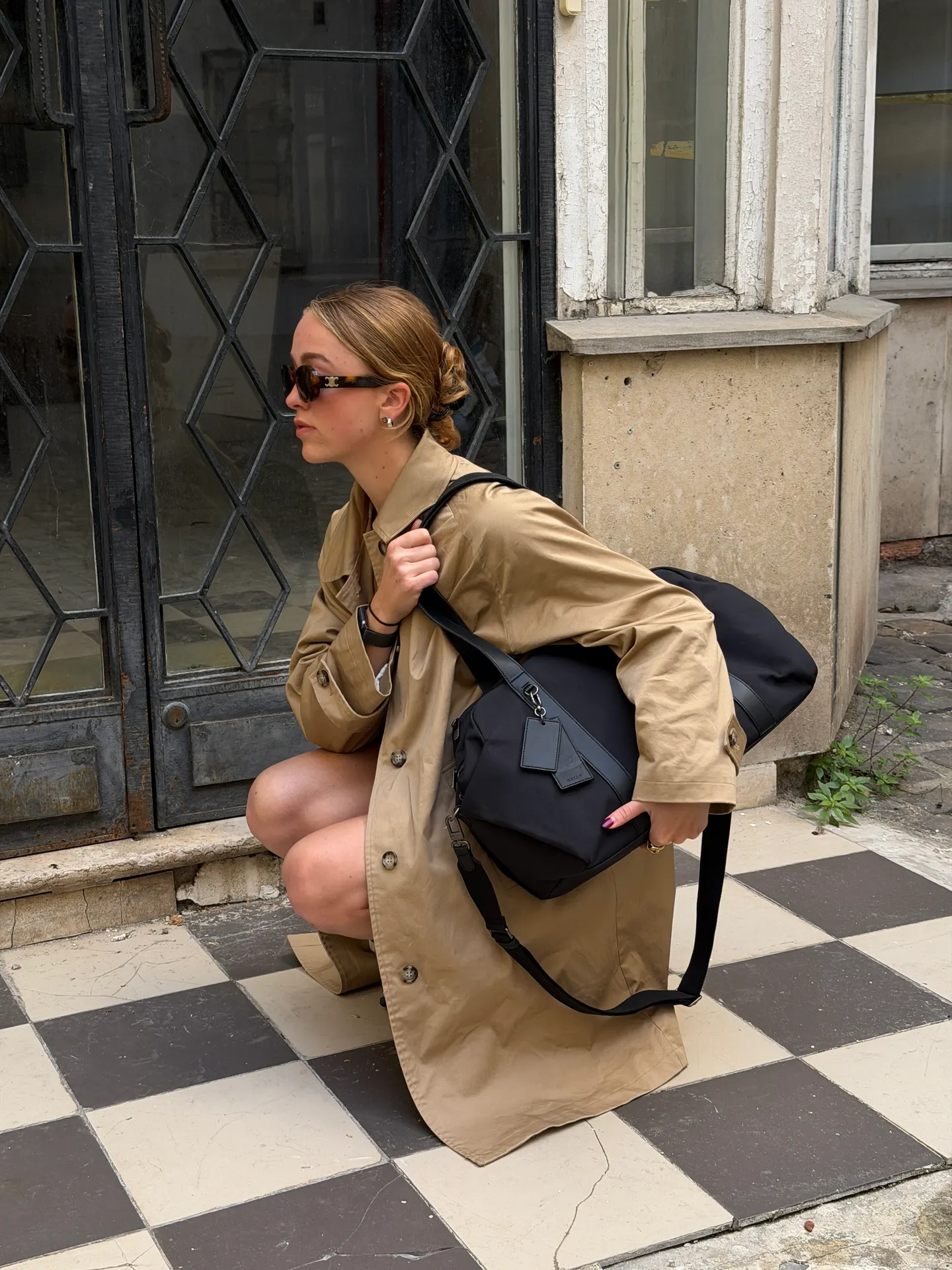 Tan trench coat, black shoulder bag, and tortoiseshell sunglasses on a checkered floor.