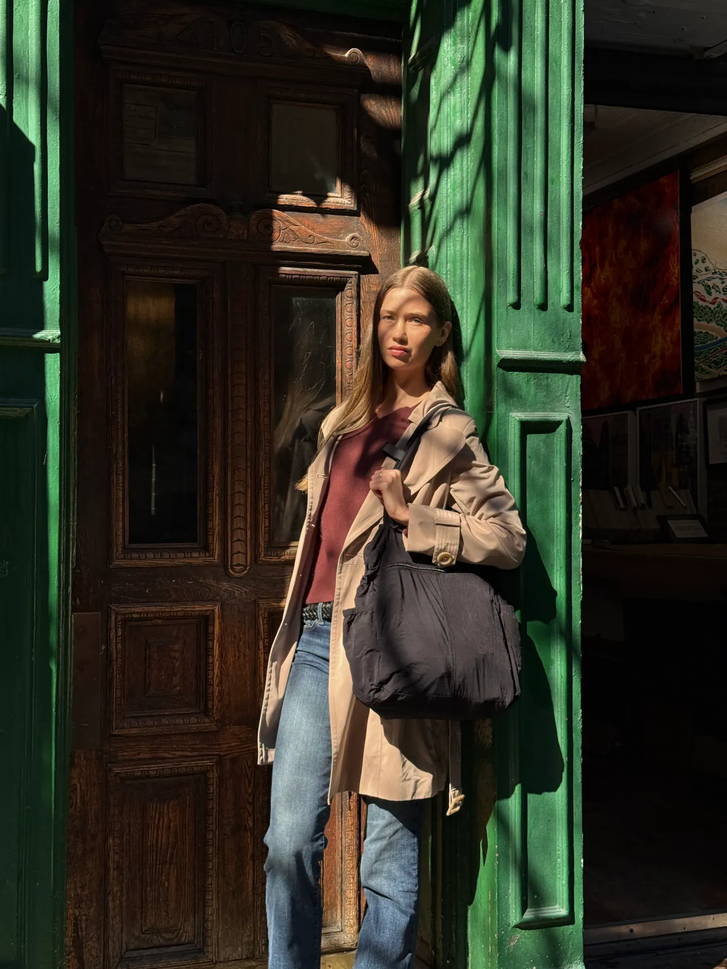 Tan trench coat with a red top, blue jeans, and a black shoulder bag next to a green pillar.