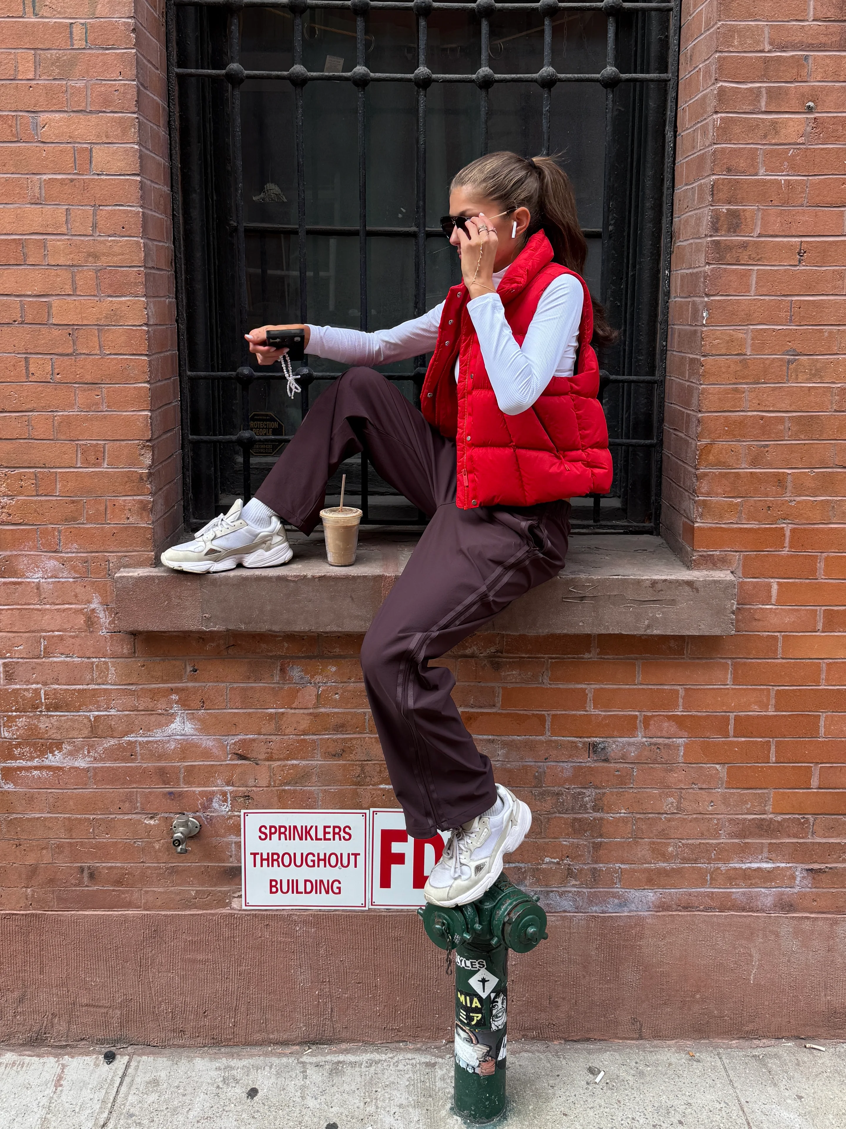 Red puffer vest, white long-sleeve, brown pants, and white sneakers against a brick wall.
