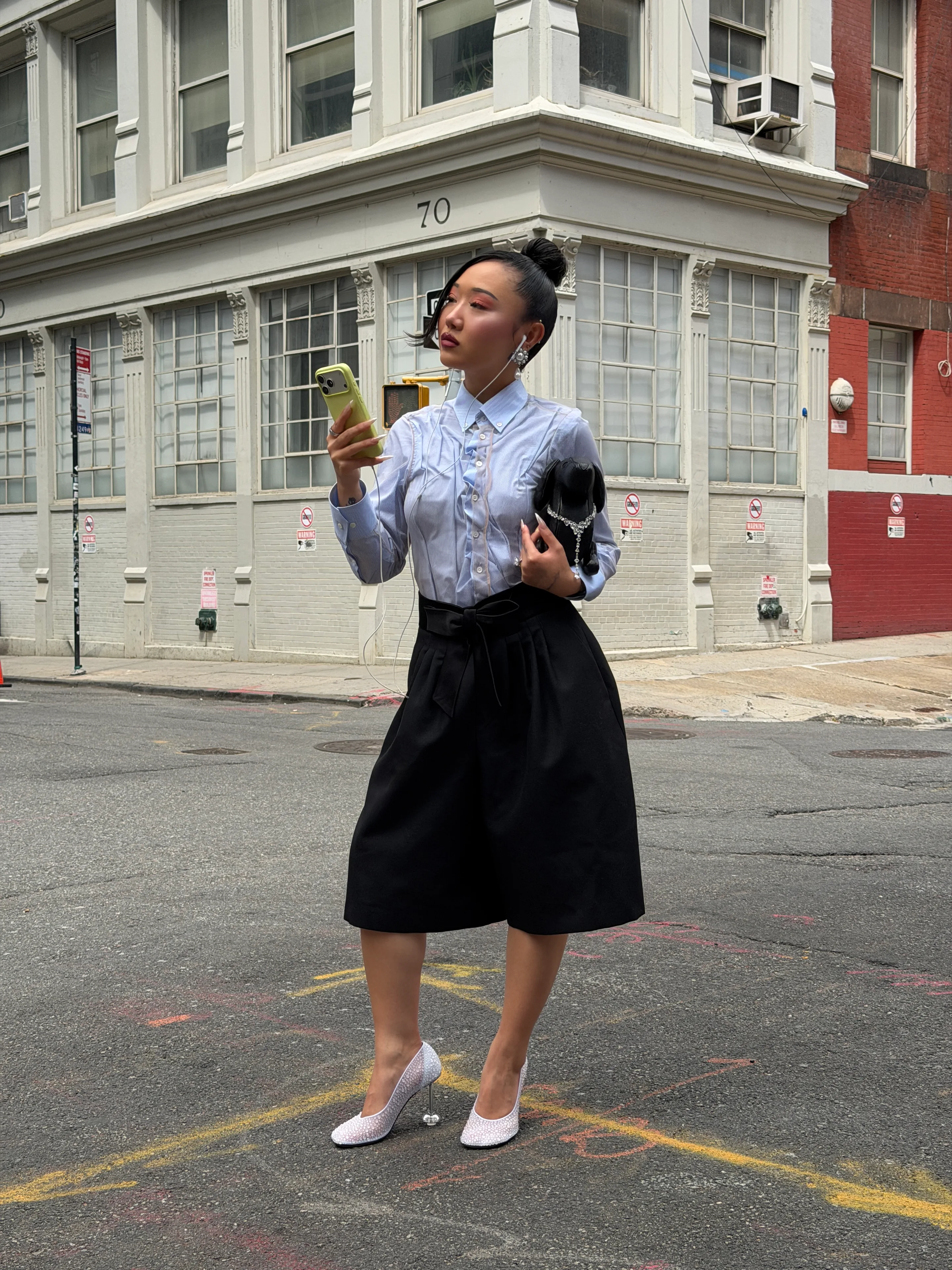 Blue pinstriped ruffle shirt, black culottes, white heels, dog bag, and earrings in a city street.