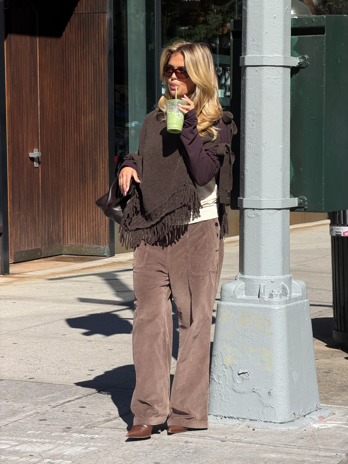Brown fringed poncho, corduroy pants, sunglasses, and green drink on a sunny street.