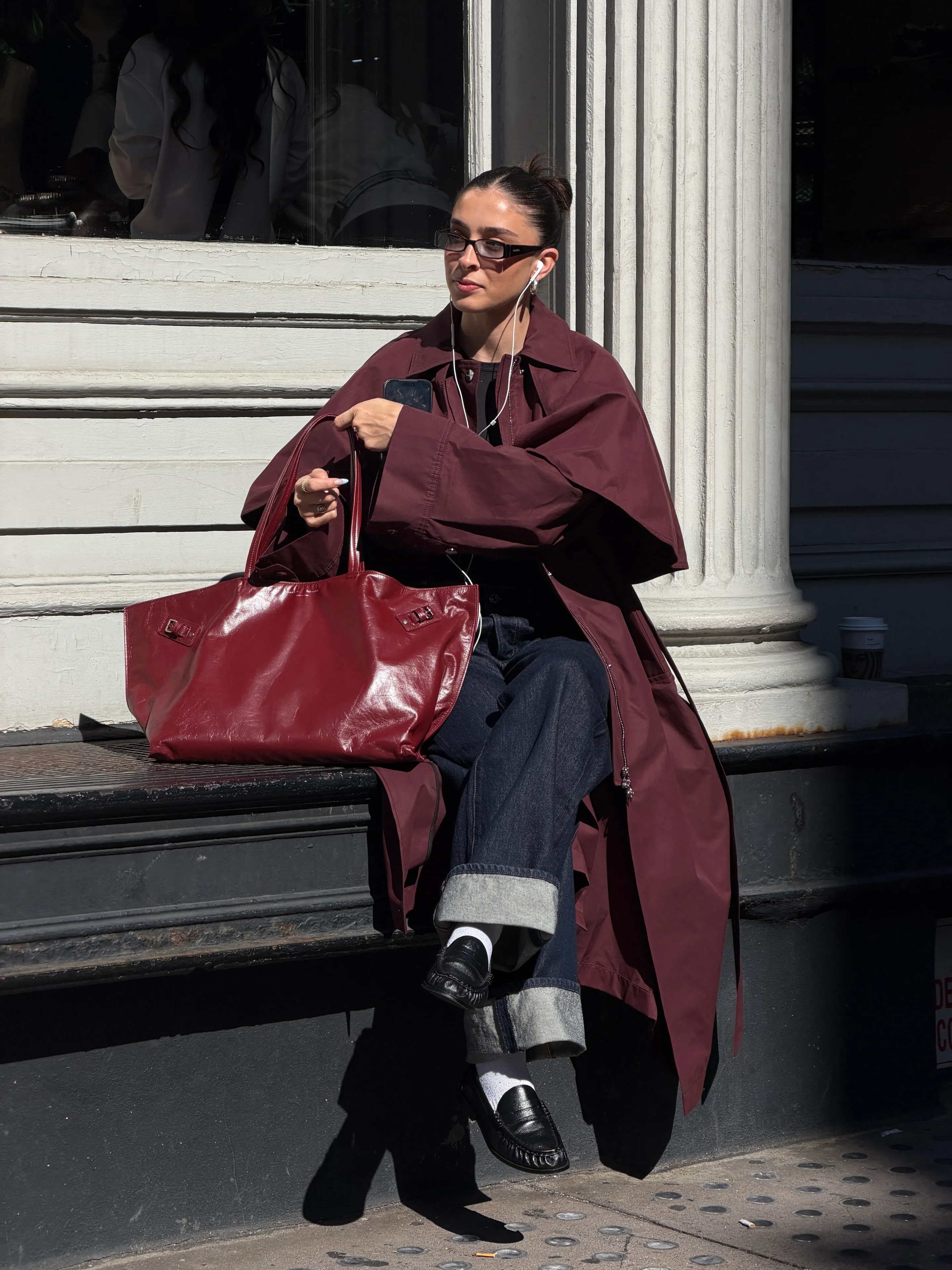 Maroon trench coat, dark wash jeans, black loafers, and red leather tote bag street style.