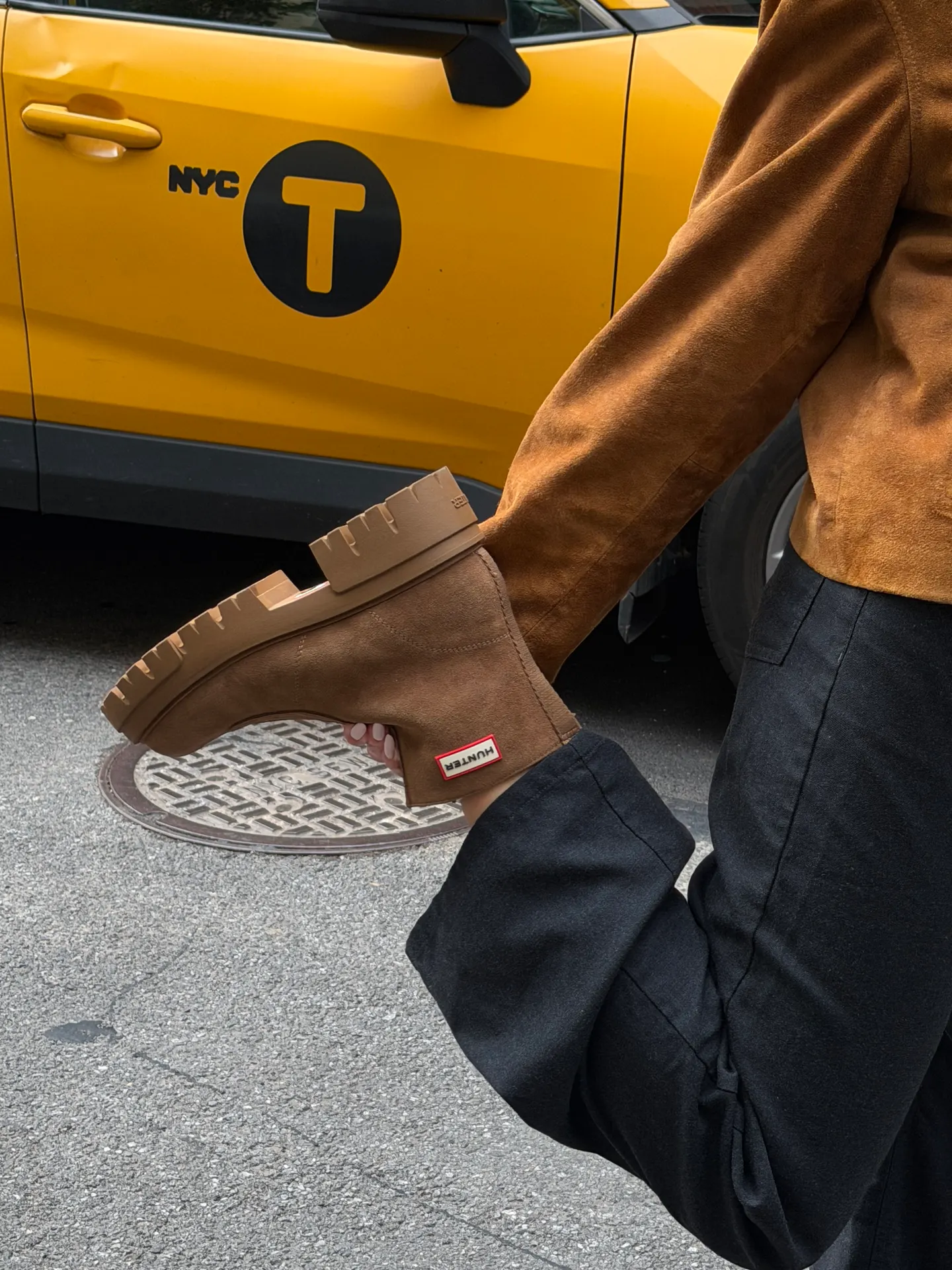 Brown suede jacket and black pants, holding a Hunter boot, with a NYC taxi in background.