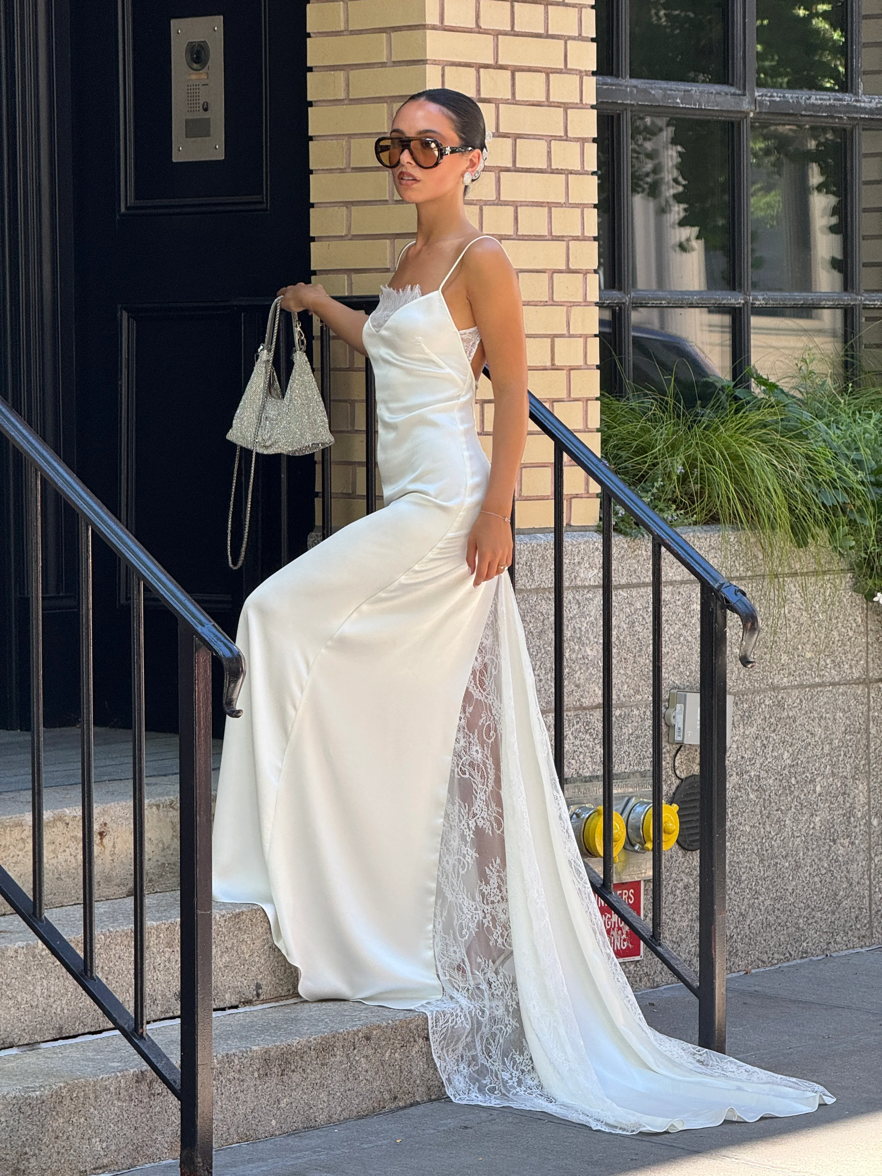 White satin slip dress with lace details, carrying a silver sequin bag and wearing sunglasses.