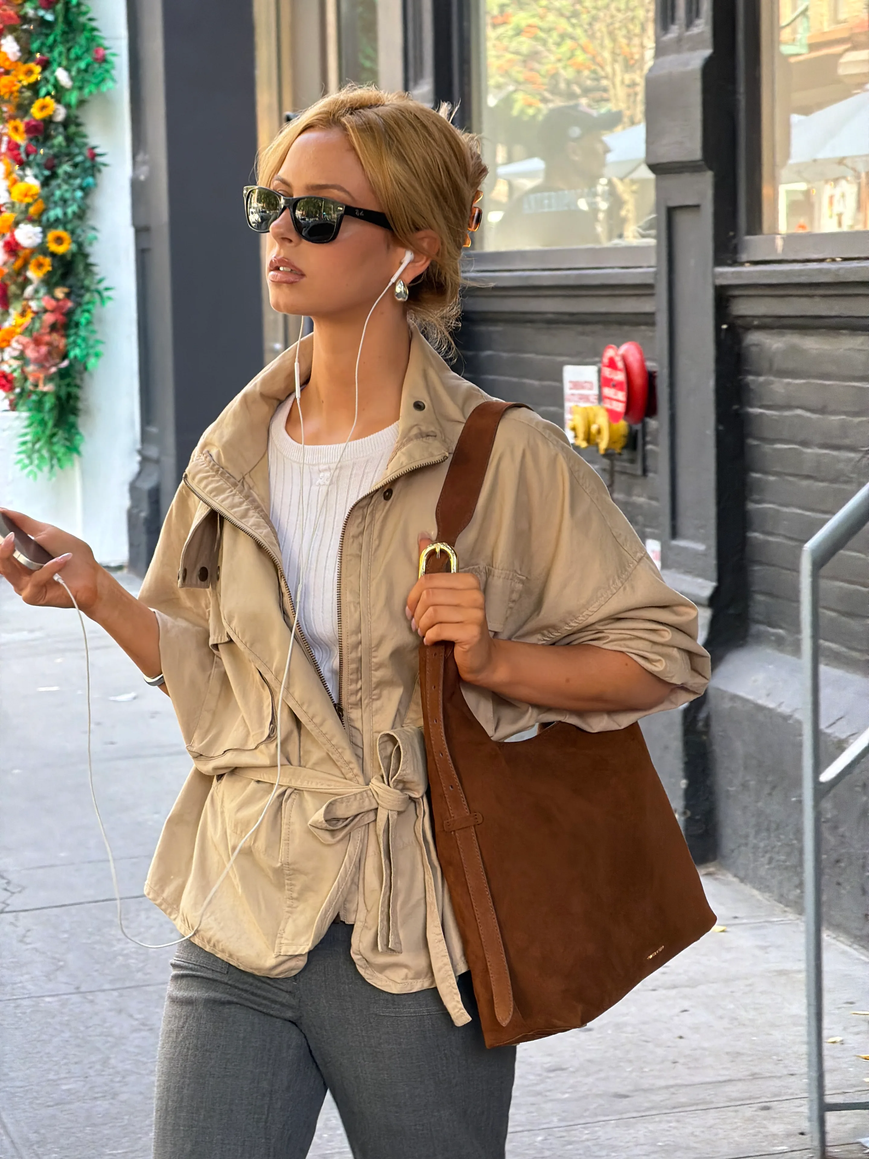 Tan utility jacket, white ribbed top, gray pants, brown suede bag, sunglasses, earbuds.