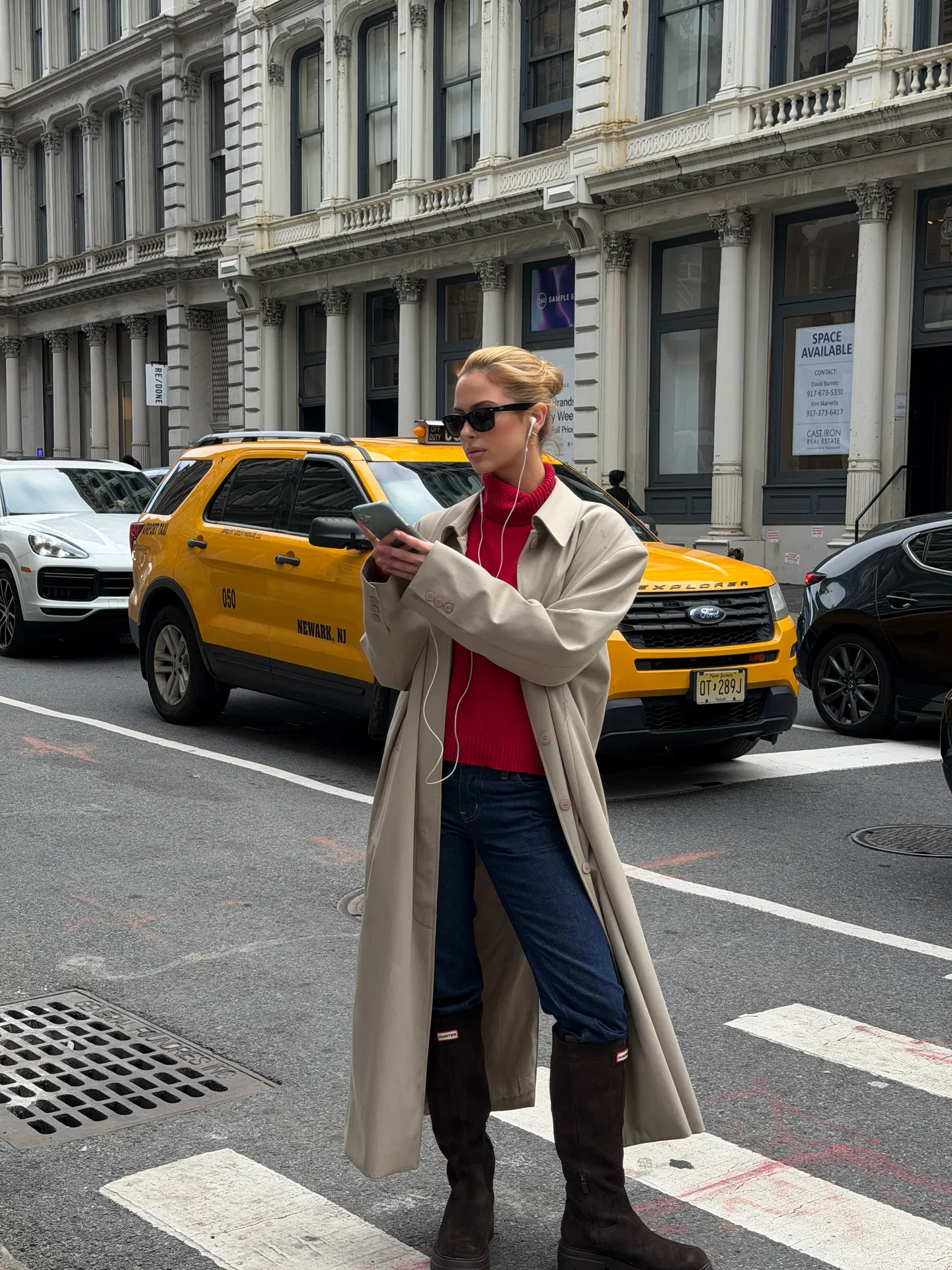 Beige trench coat with a red turtleneck, blue jeans, and brown Hunter boots on a city street.