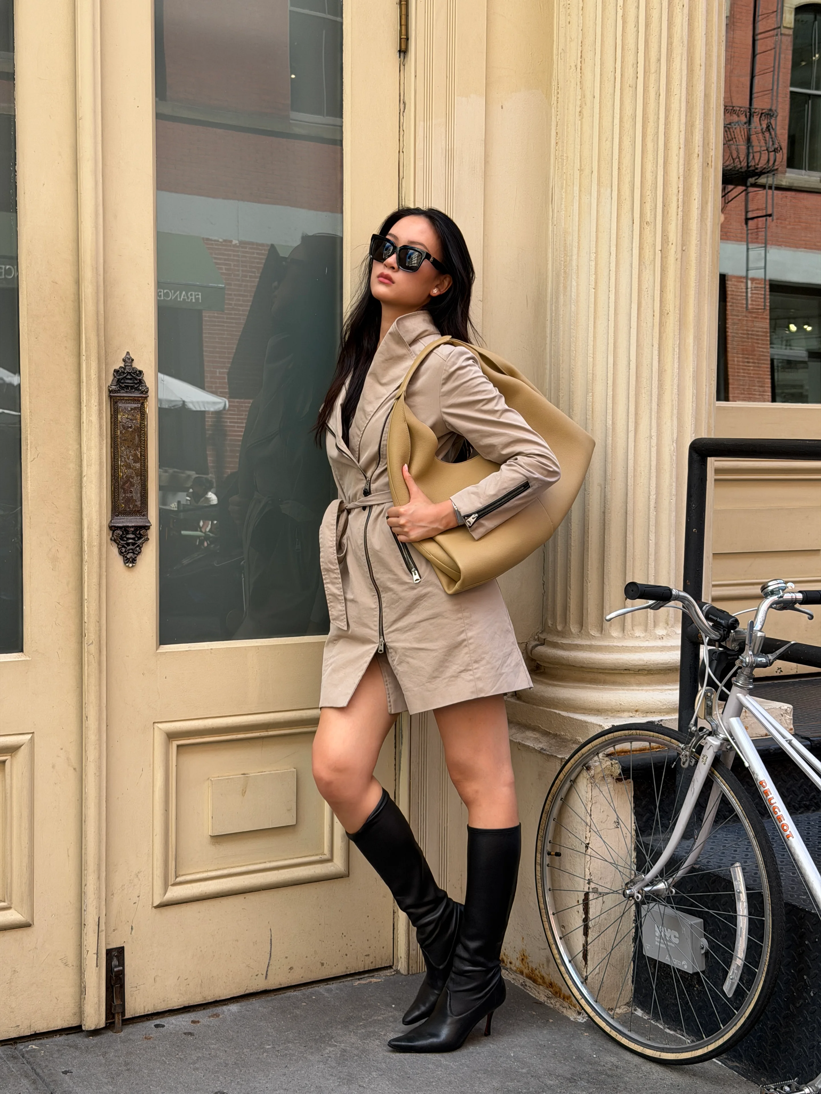 Beige trench coat dress with black boots, oversized tan bag, and sunglasses.