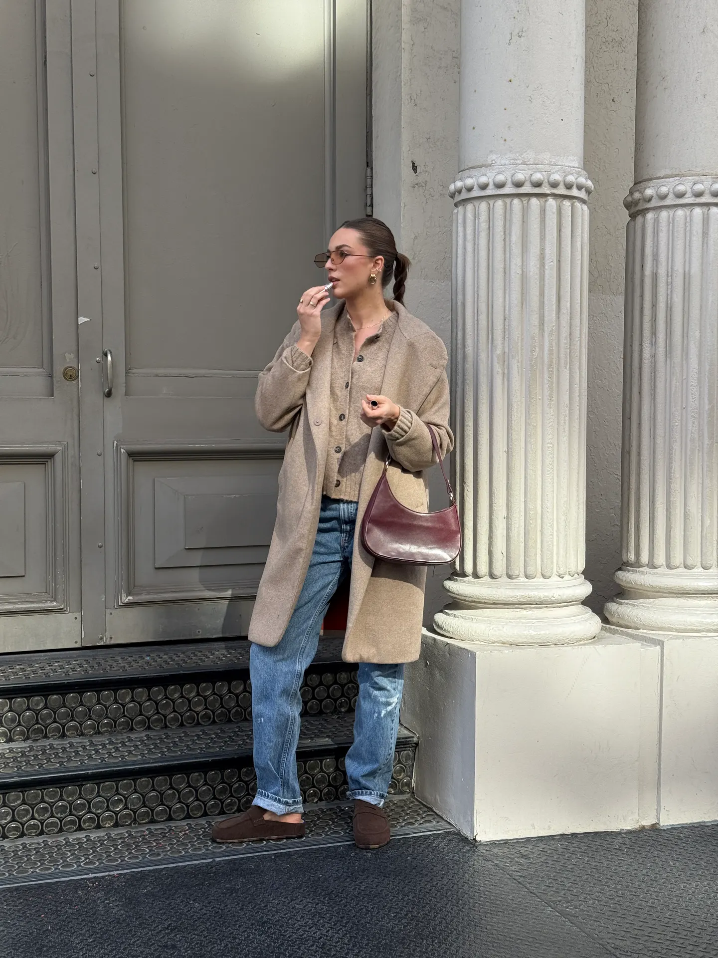 Tan long coat, matching cardigan, blue jeans, and brown mules with a maroon shoulder bag.
