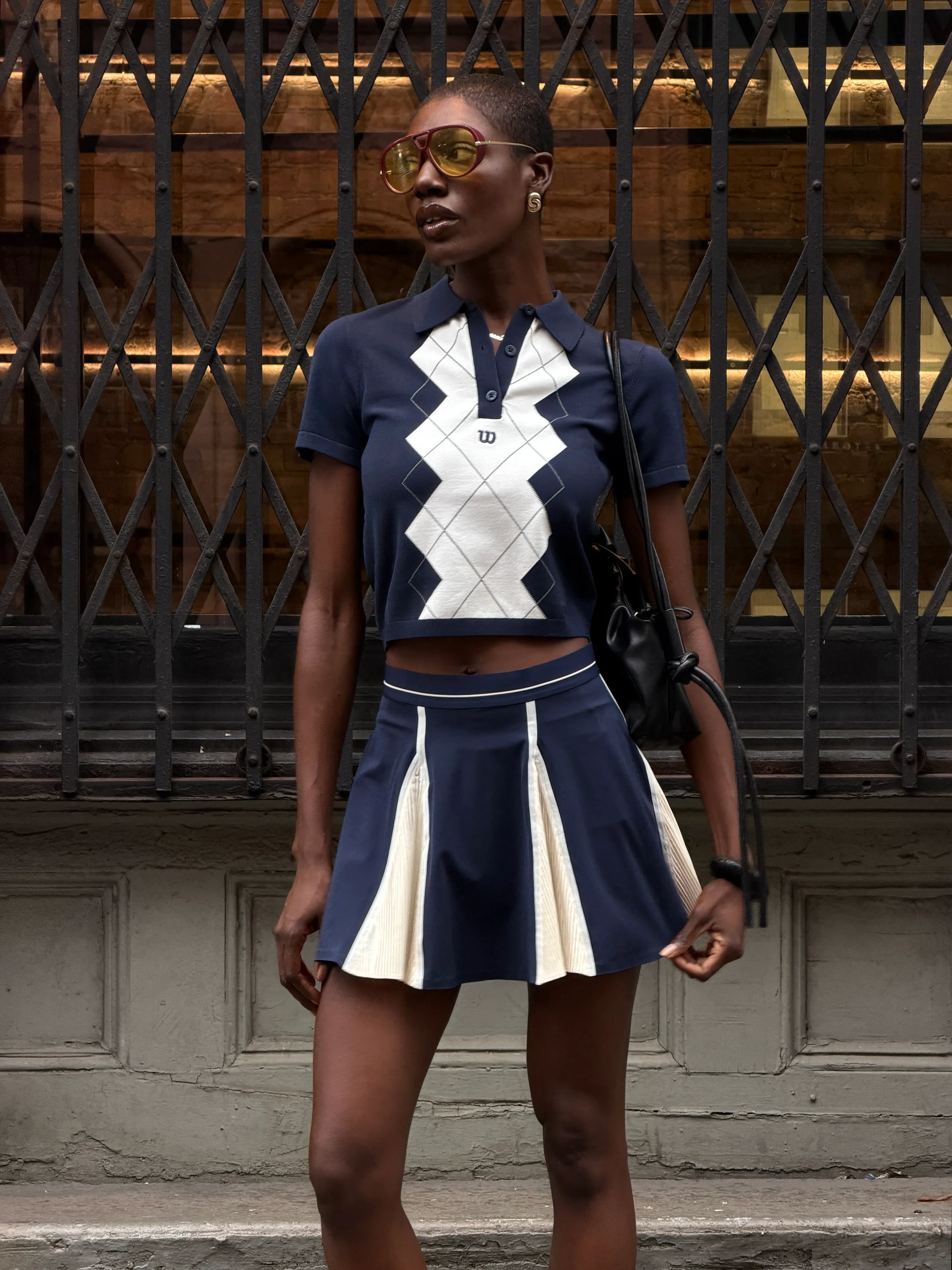 Navy and white argyle polo top with matching pleated skirt, aviator sunglasses, and black bag.