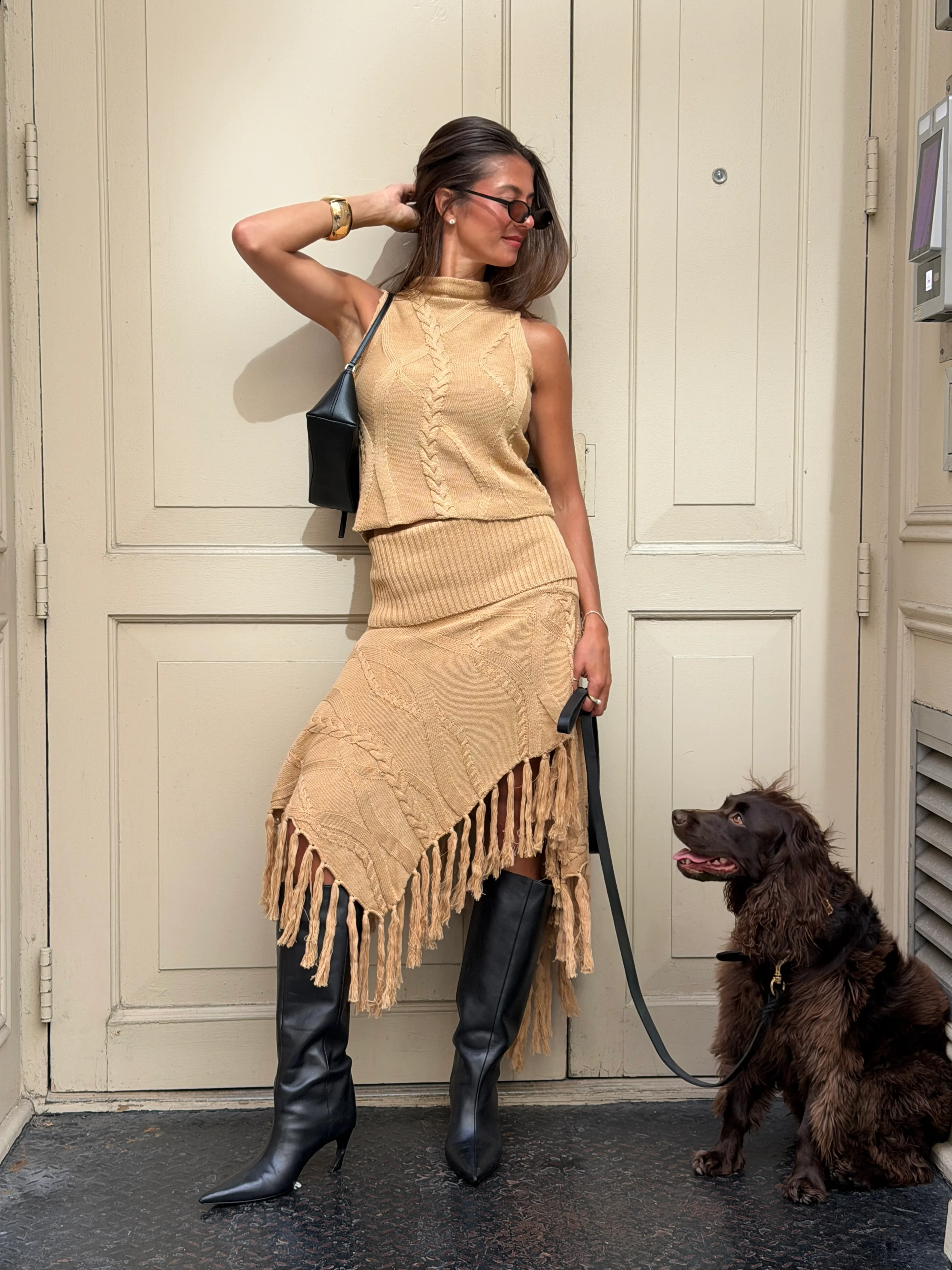 Tan cable knit top and fringed skirt set with black boots and bag, beside a brown dog.
